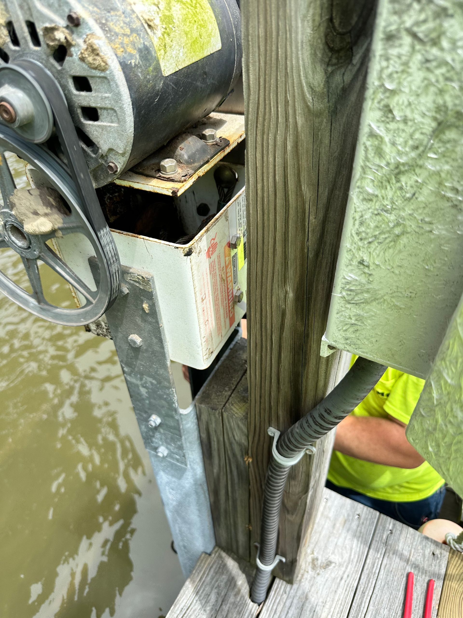Motor, pulley, and electrical box mounted on a wooden post over water. A person in yellow is partially visible.