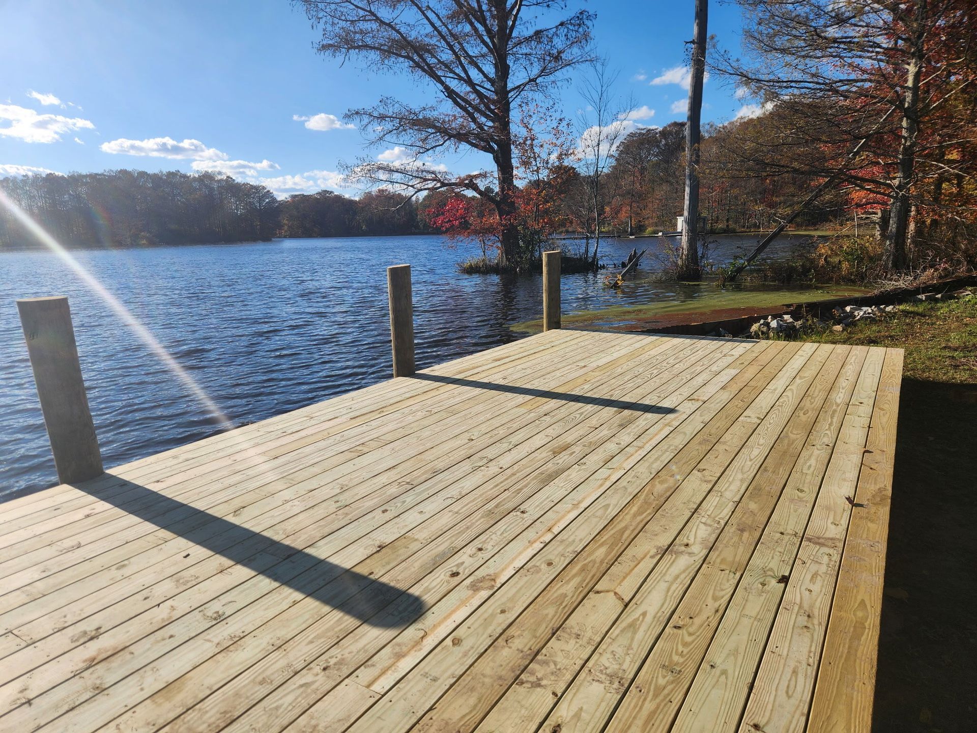 A wooden dock extends into a lake on a sunny day, with trees and a clear blue sky in the background.