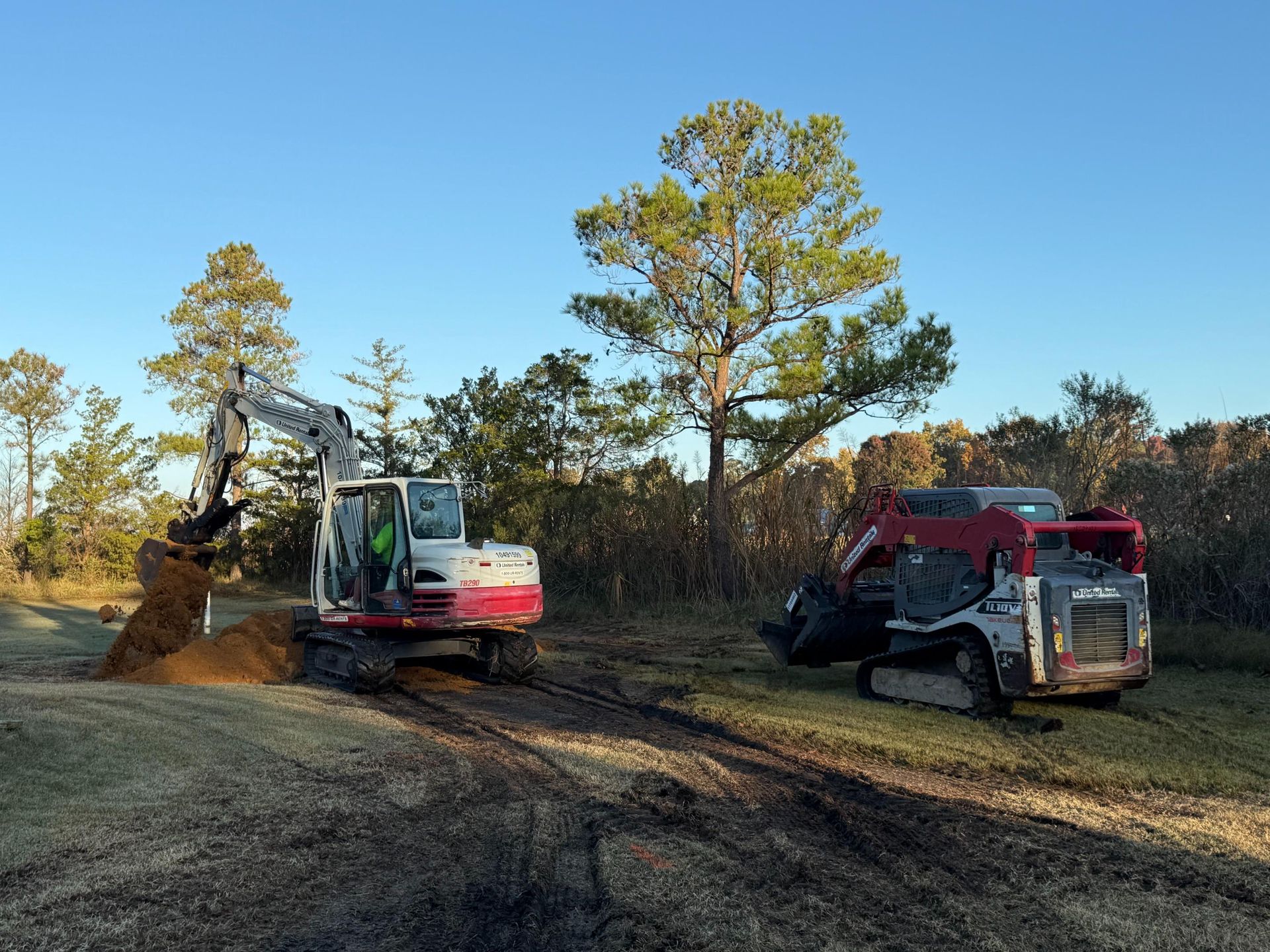 An excavator and skid steer on a grassy field, digging in front of trees under a blue sky.