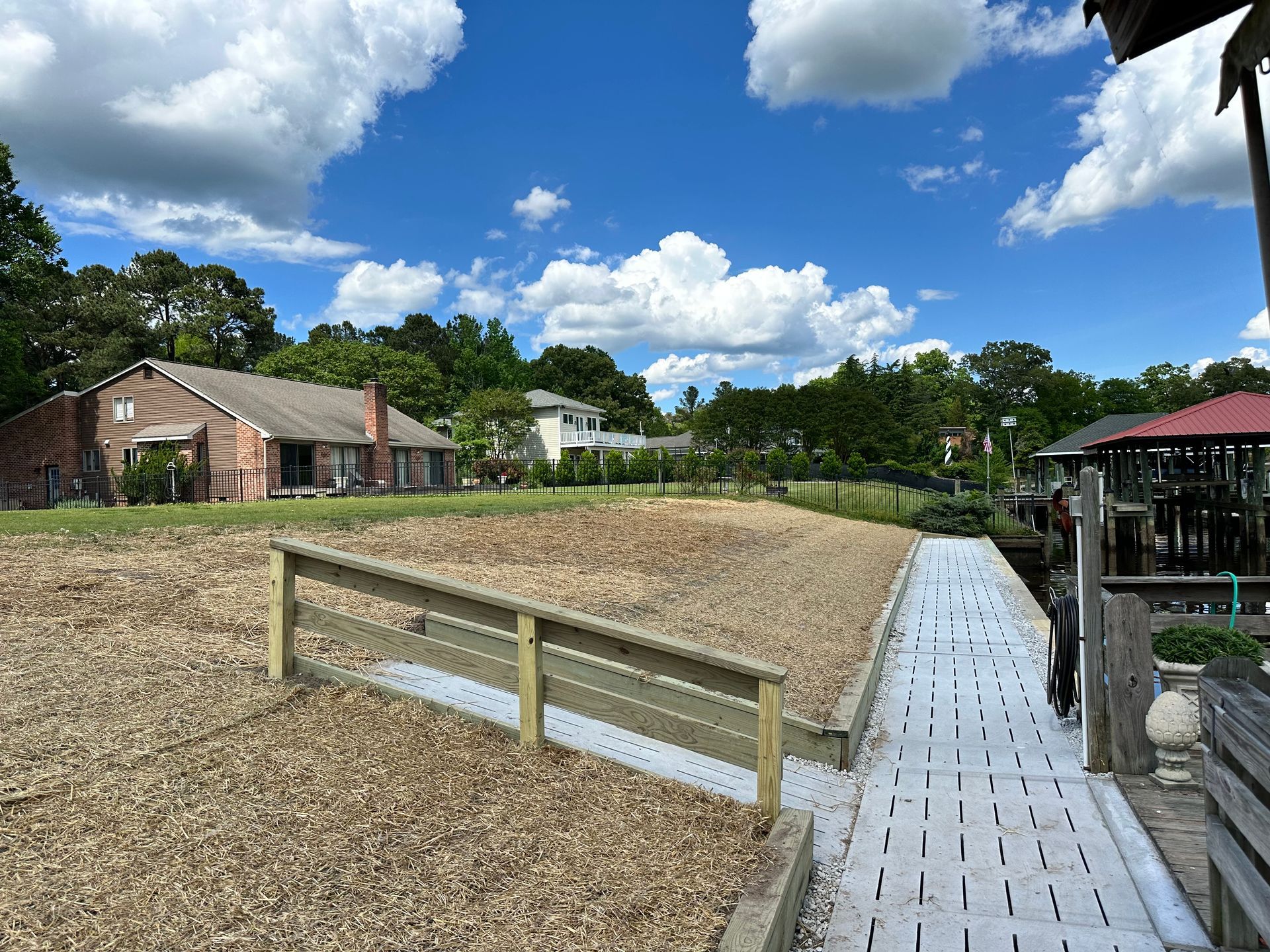 Wooden dock leads to a gravel area with a house and trees in the background under a blue sky.