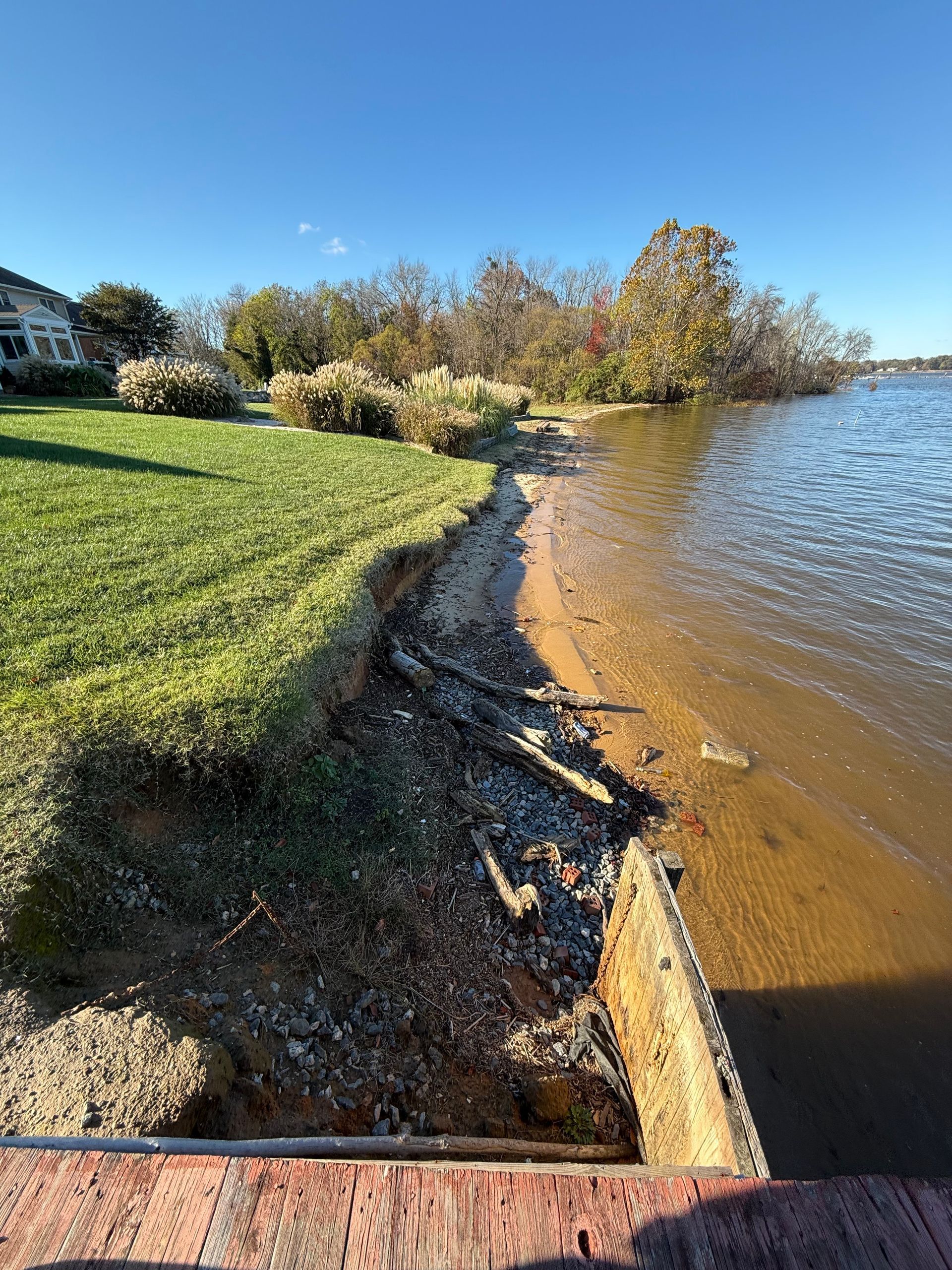 Eroding shoreline, featuring green grass, sandy bank, and water under a bright blue sky.