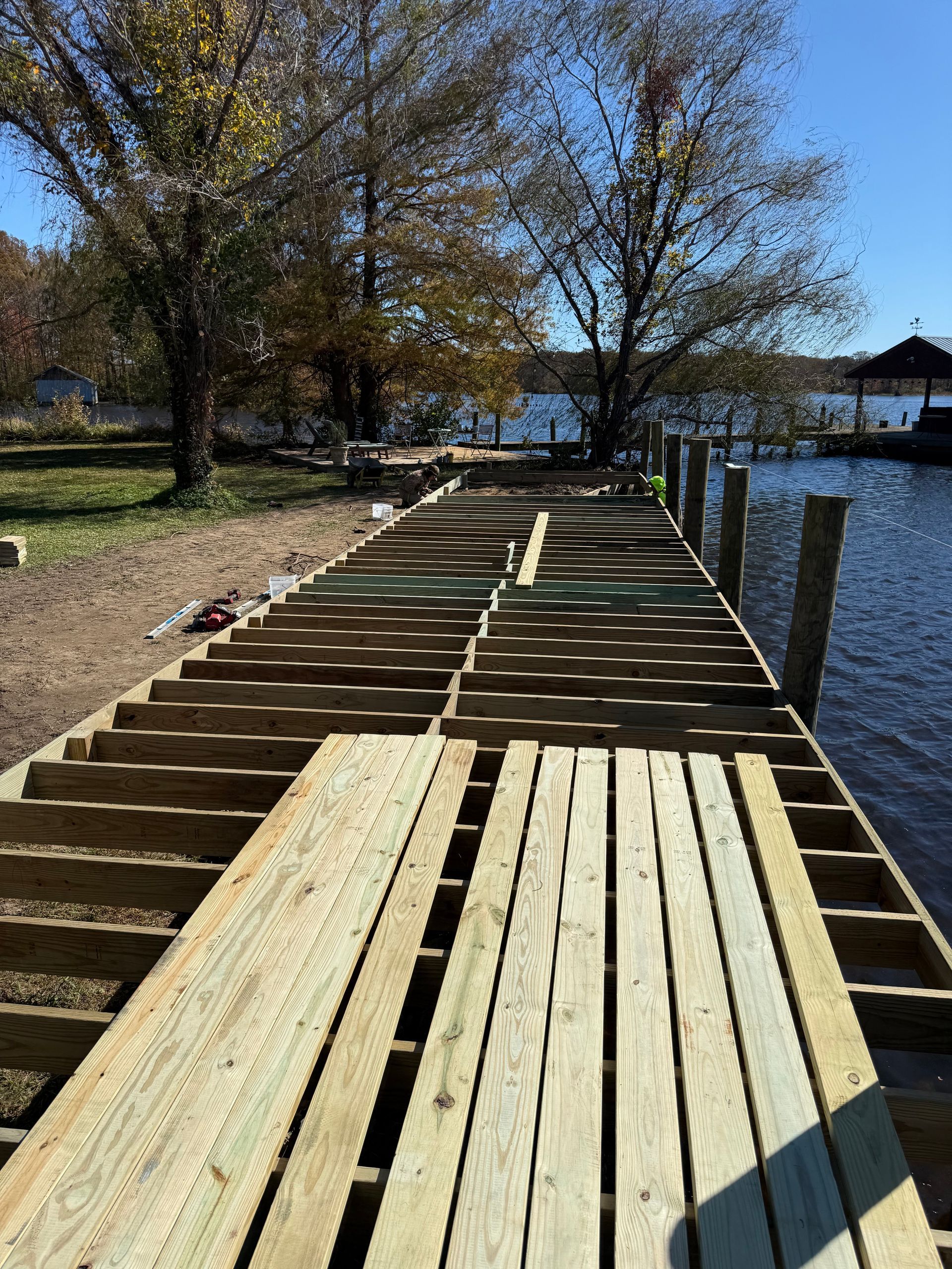 A wooden dock under construction with planks being laid over exposed framing beams, extending into a calm lake.