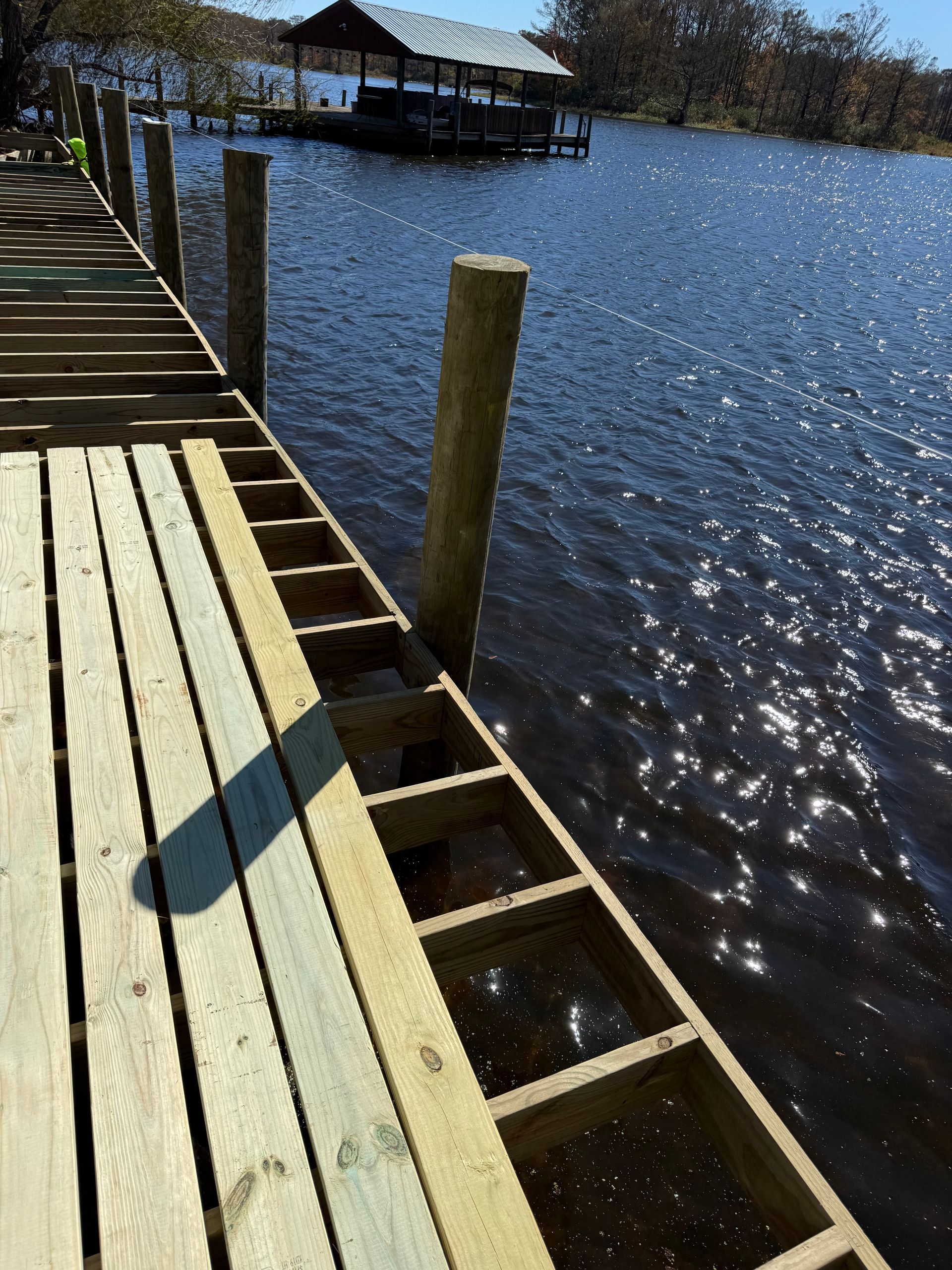 A wooden dock under construction on a sunny lake, with an unfinished plank section and a boat slip in the distance.