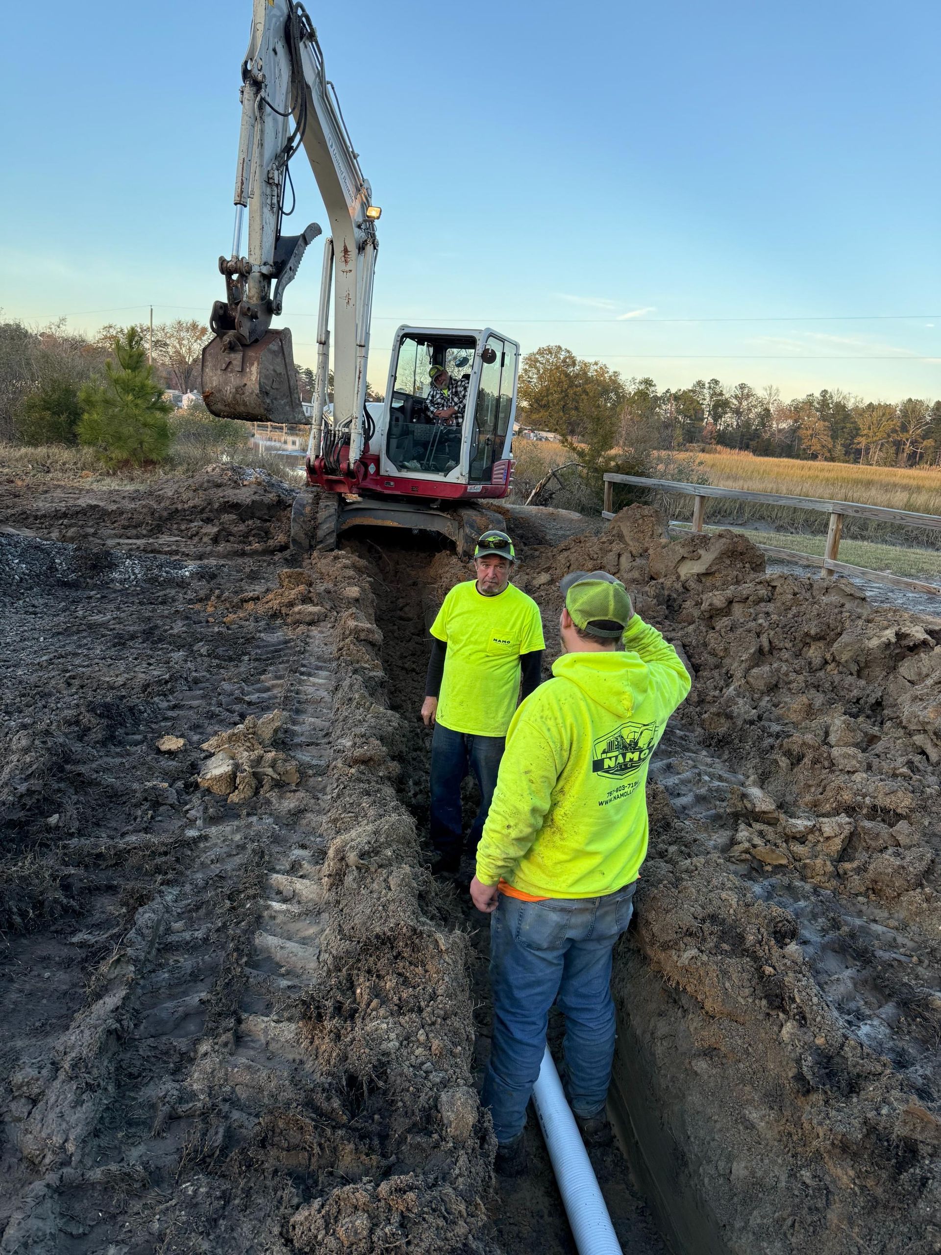 Two workers in neon shirts inspect a pipe in a ditch, excavator in background.