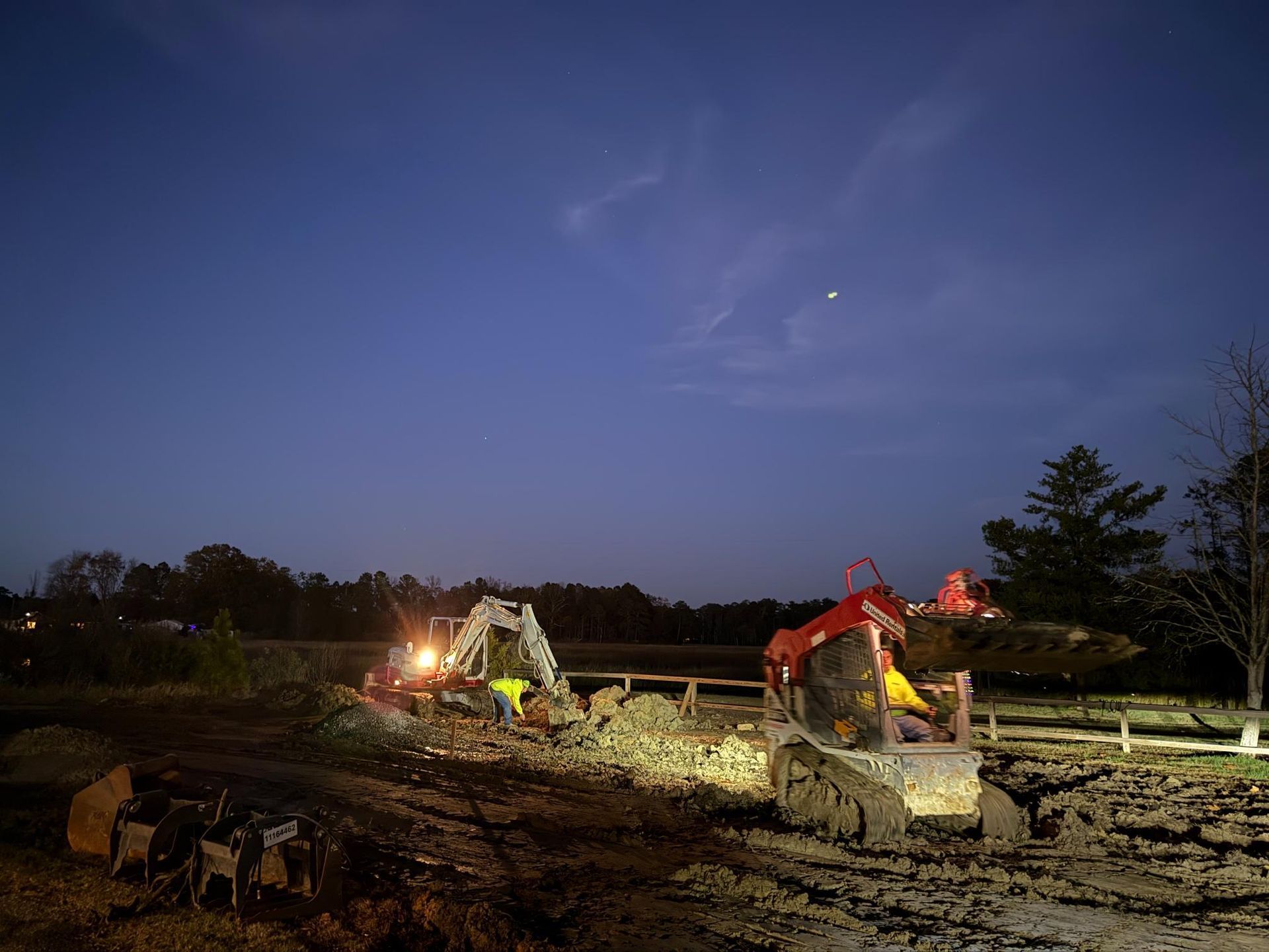 Construction site at dusk, two excavators lit up, digging in dirt under a blue sky.