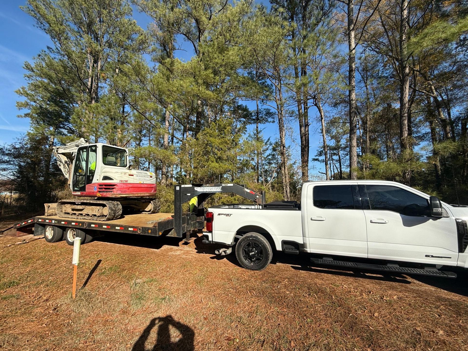 White truck towing an excavator on a trailer on a dirt road. Trees in the background, sunny day.
