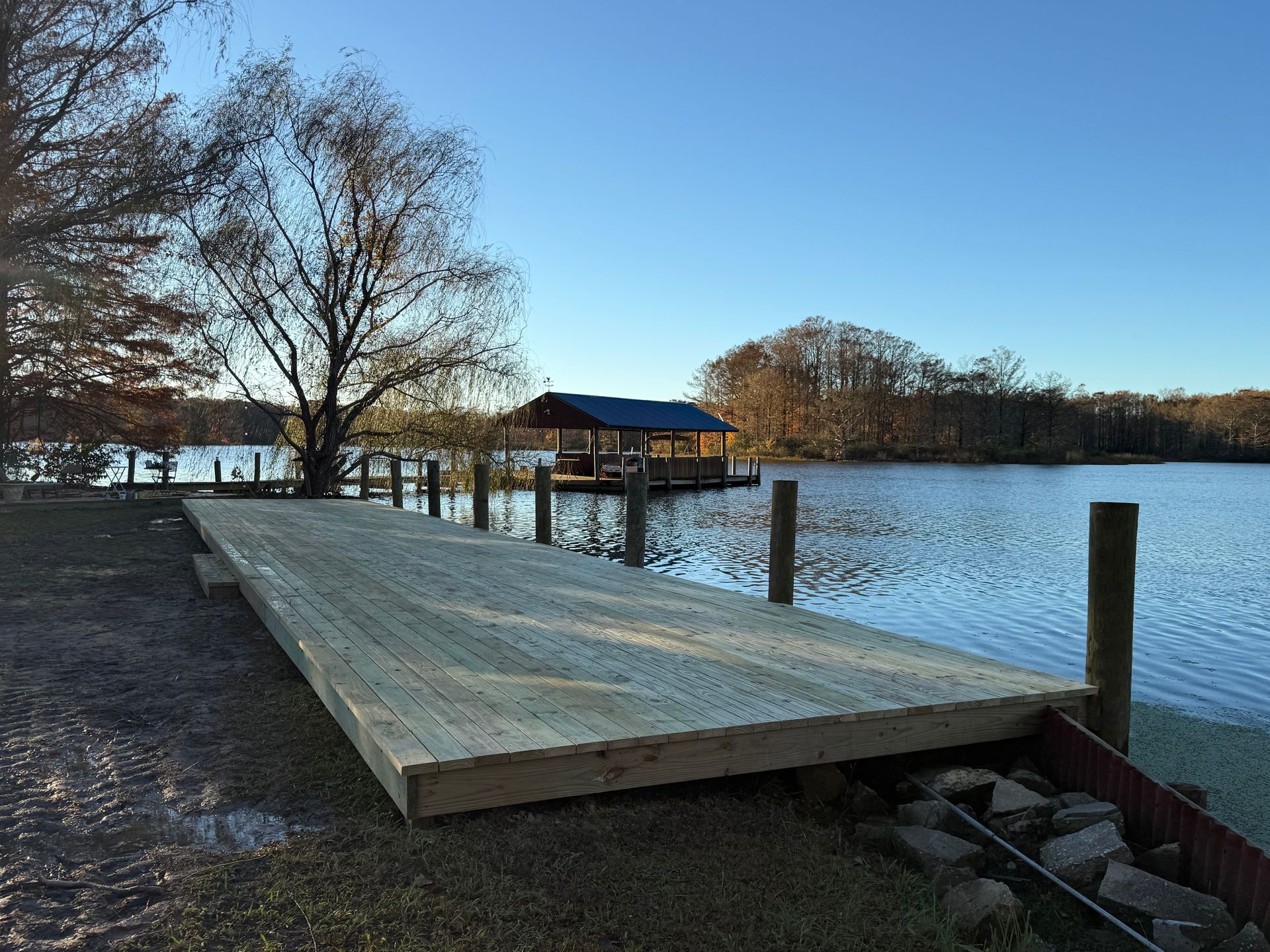 A newly built wooden deck extends from a grassy bank into a calm lake under a clear blue sky, with a boathouse beyond.