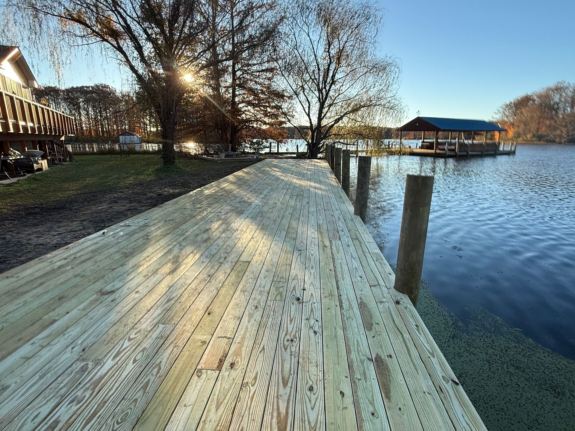 Newly built wooden dock extending into a lake, with sunlit trees and a boathouse in the background.