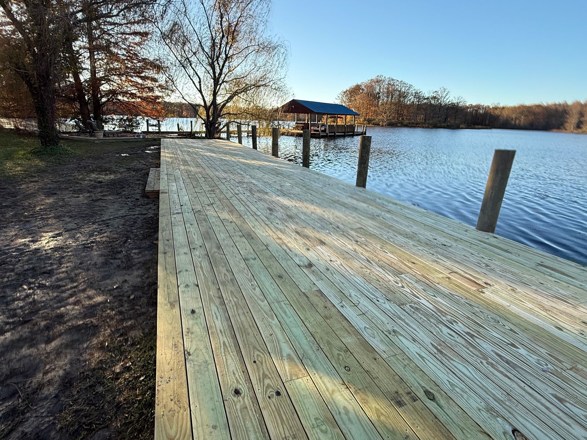 A newly built wooden boat dock extending into a calm lake surrounded by trees under a clear blue sky.