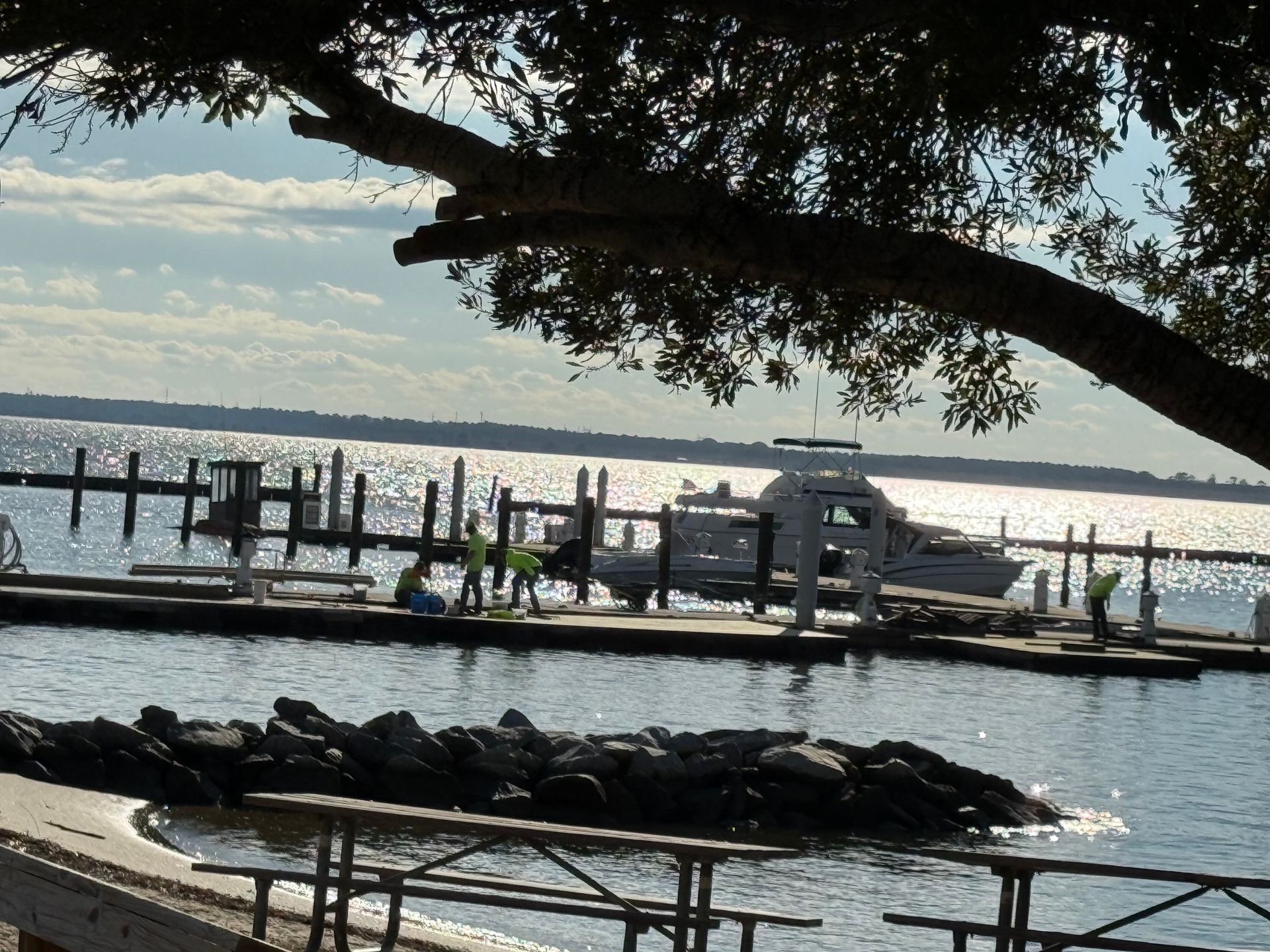 A boat docked at a pier, with people, rocks, and picnic tables in the foreground. Sun is shining on water.