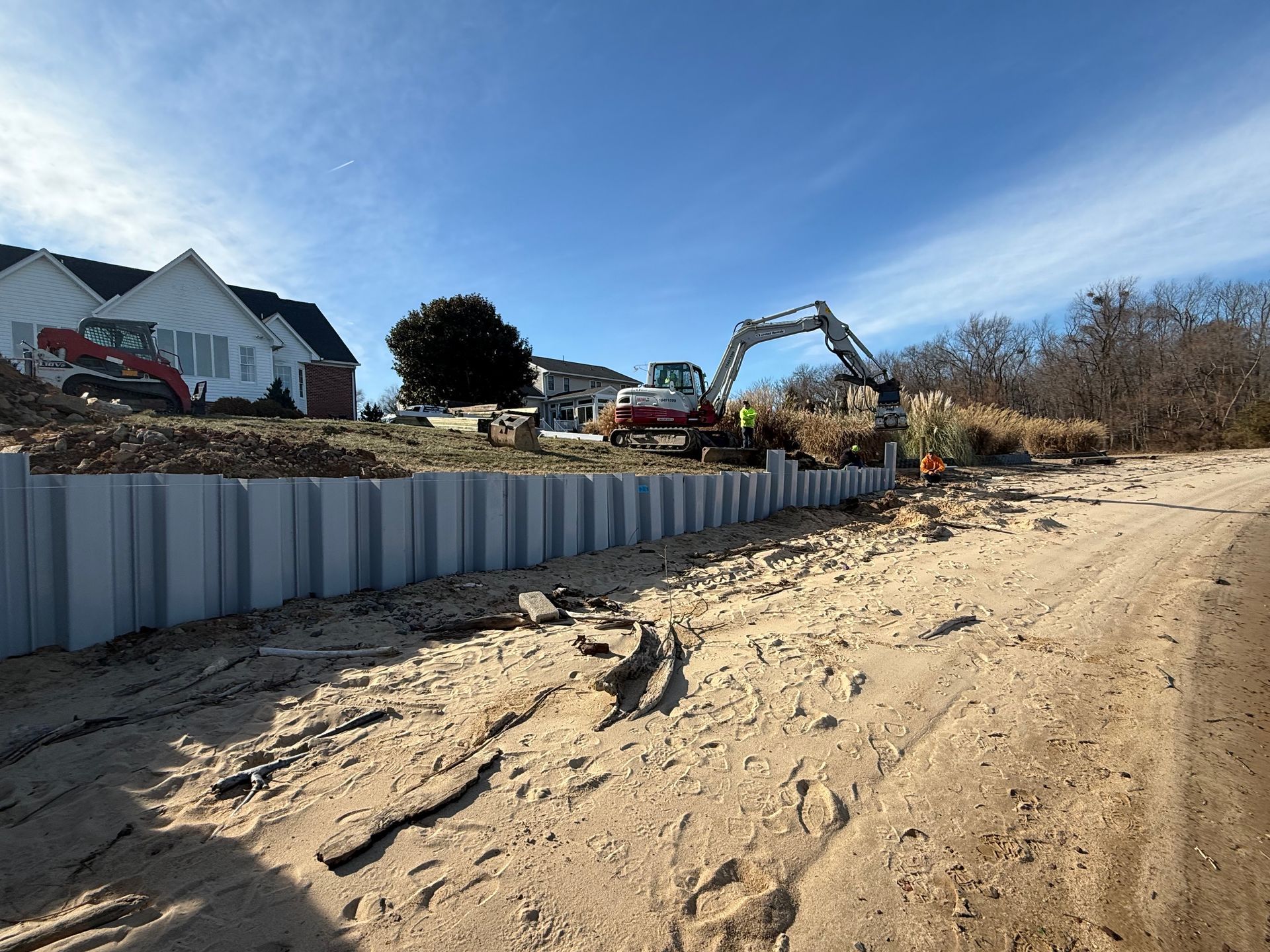 Construction site with a retaining wall, excavator, and houses on a sunny day.