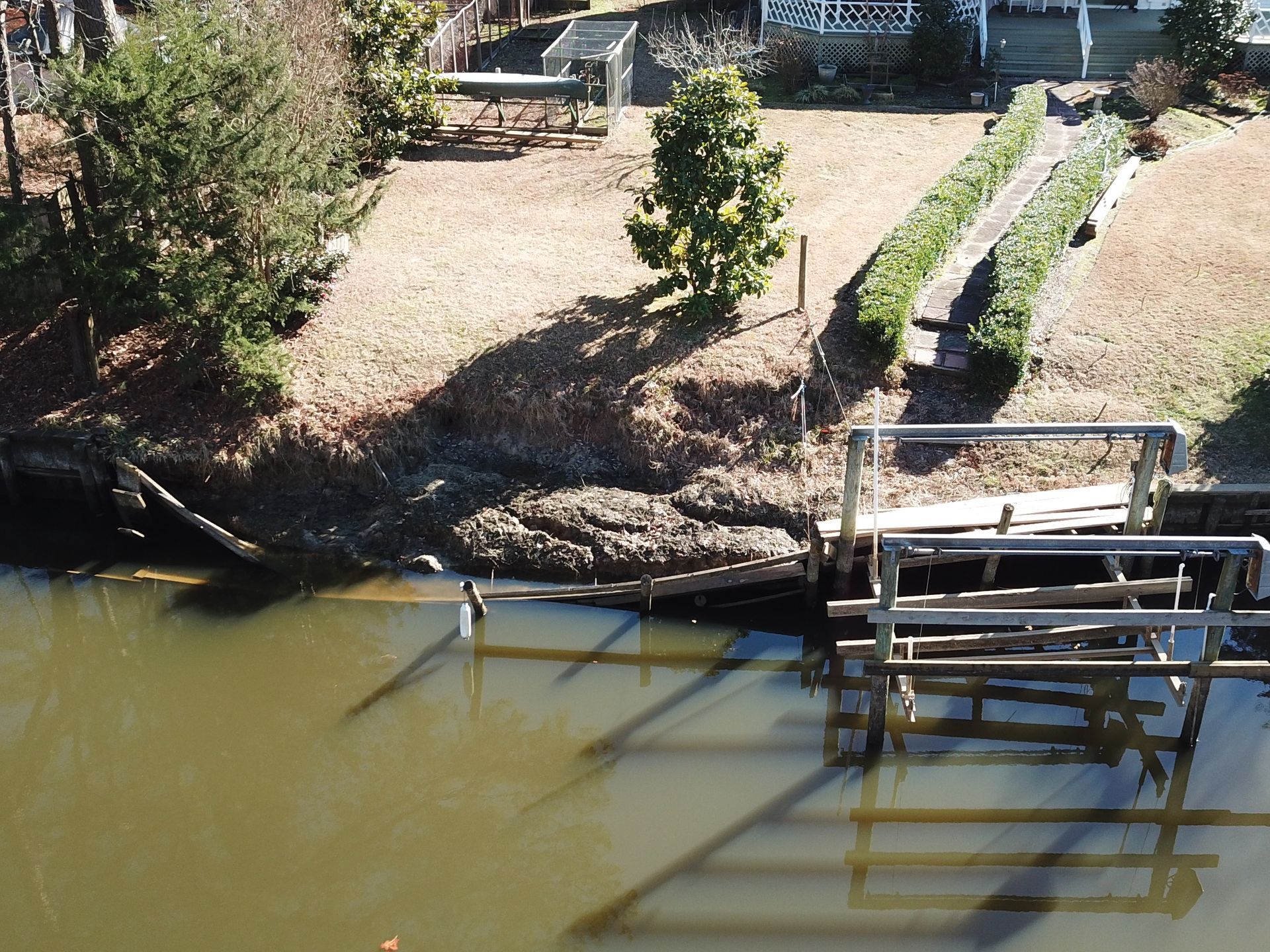 An elevated view of a brown waterway featuring a damaged wooden dock structure along a grassy, sloped shoreline.