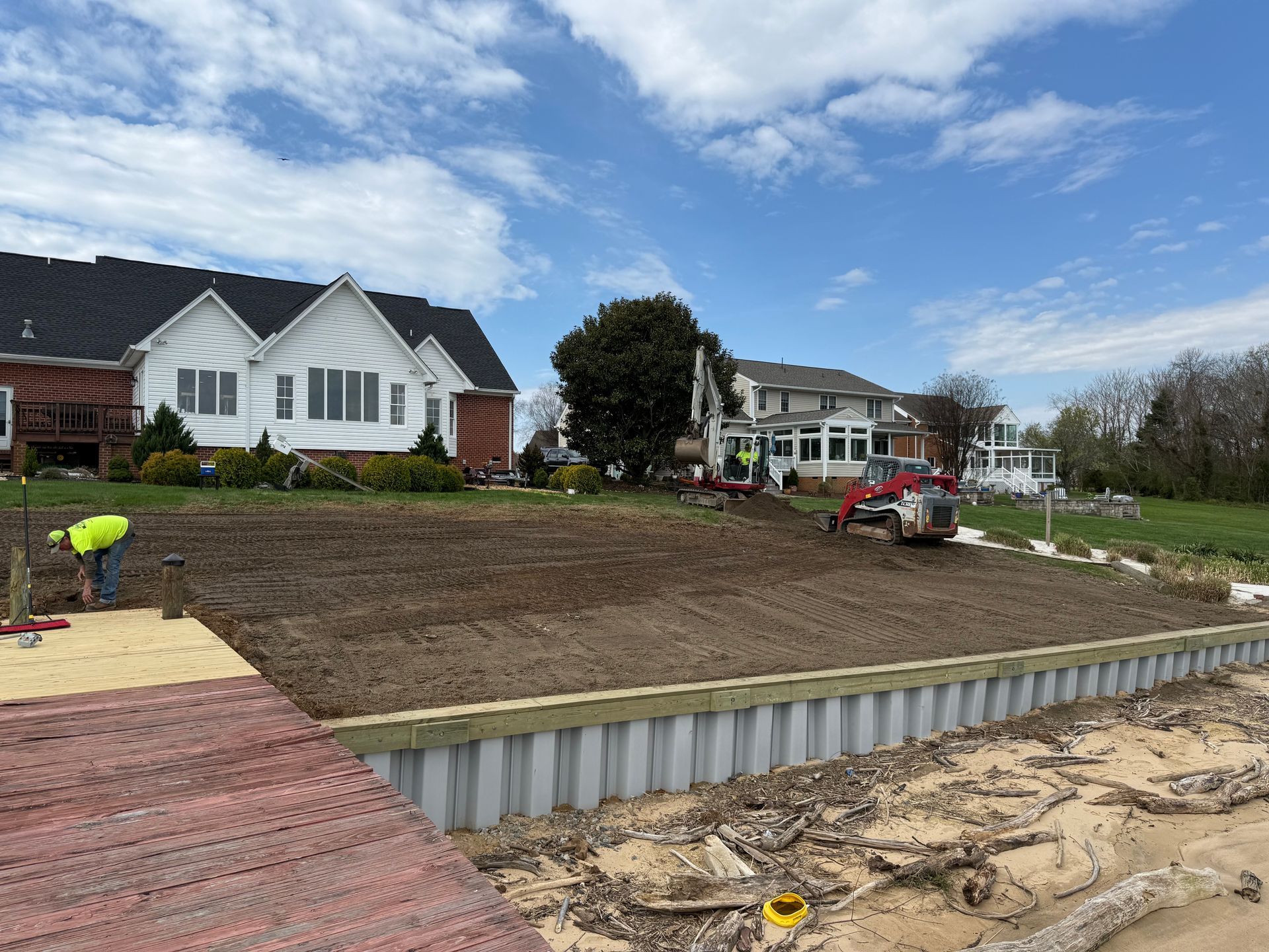 Construction site near water with houses. Workers, a bobcat, and a retaining wall on a beach.