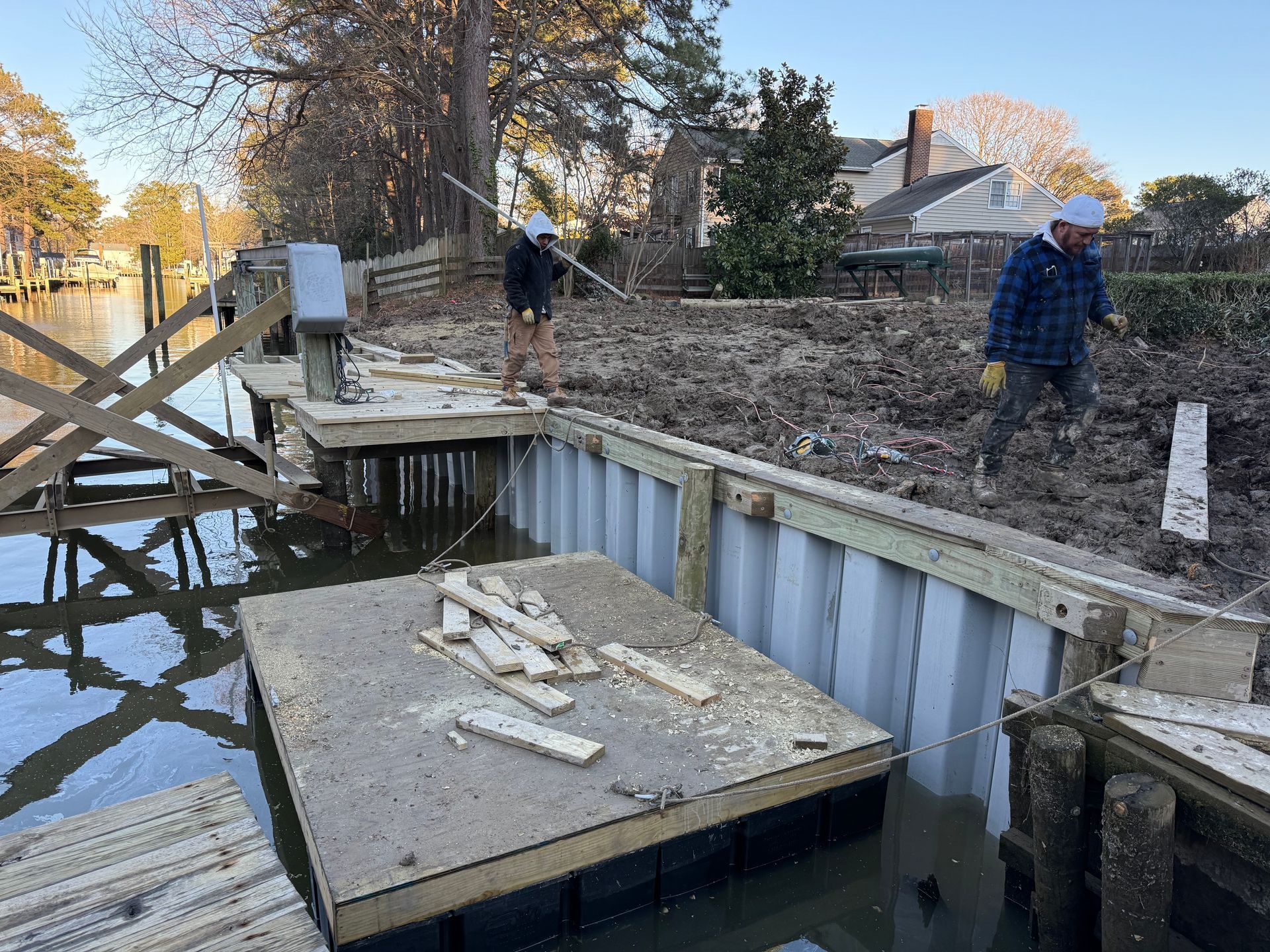 Two workers build a bulkhead along a waterfront property, with a floating platform and new grey wall panels installed.