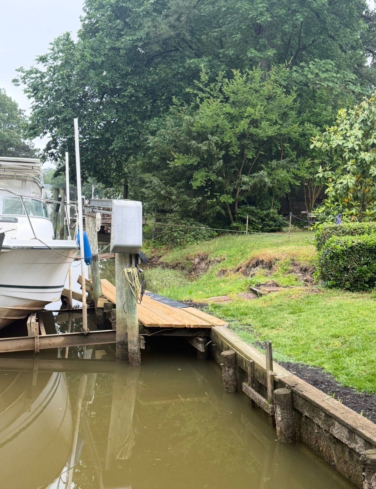 A boat moored at a wooden pier alongside a grassy bank with a bulkhead and dense trees in the background.