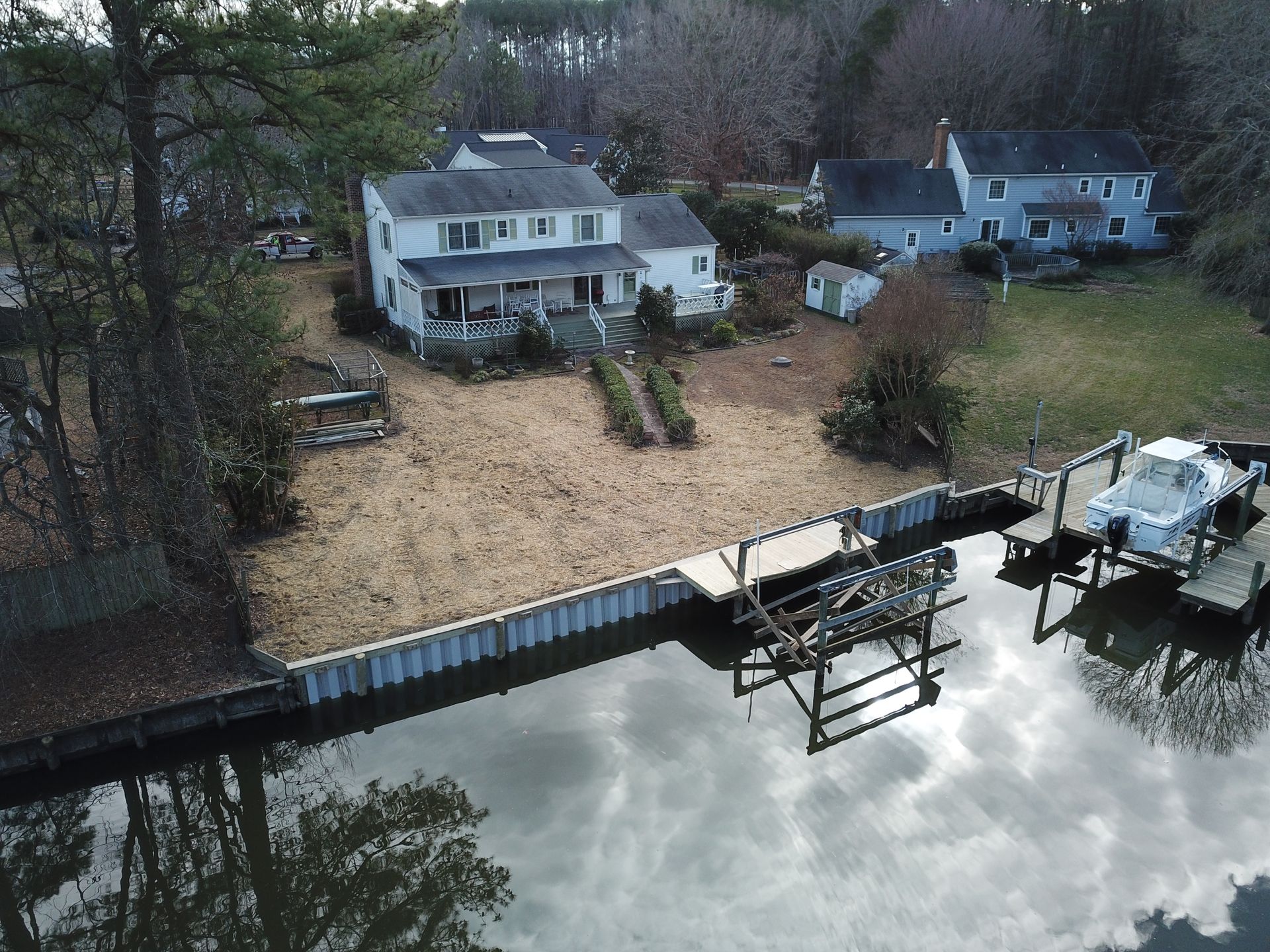 An aerial view of a waterfront property with a white house, a bulkheaded yard, and a boat lift with a boat in the water.