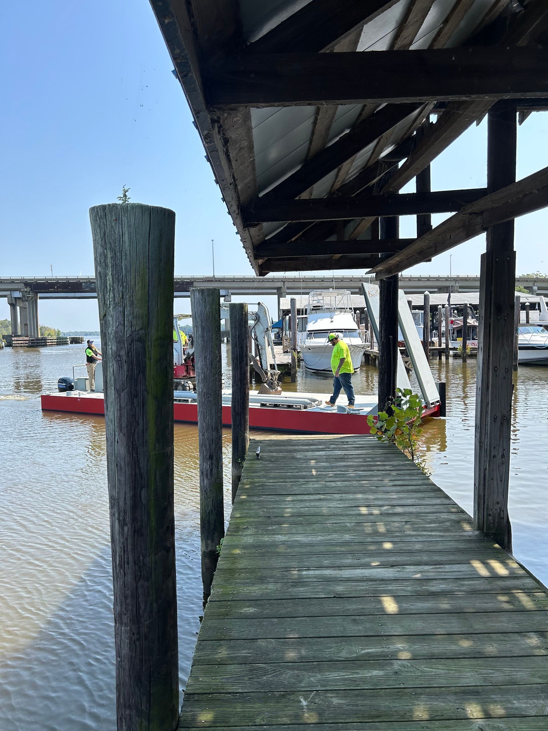 Wooden dock with a weathered roof, looking out at boats and a bridge on a sunny day.