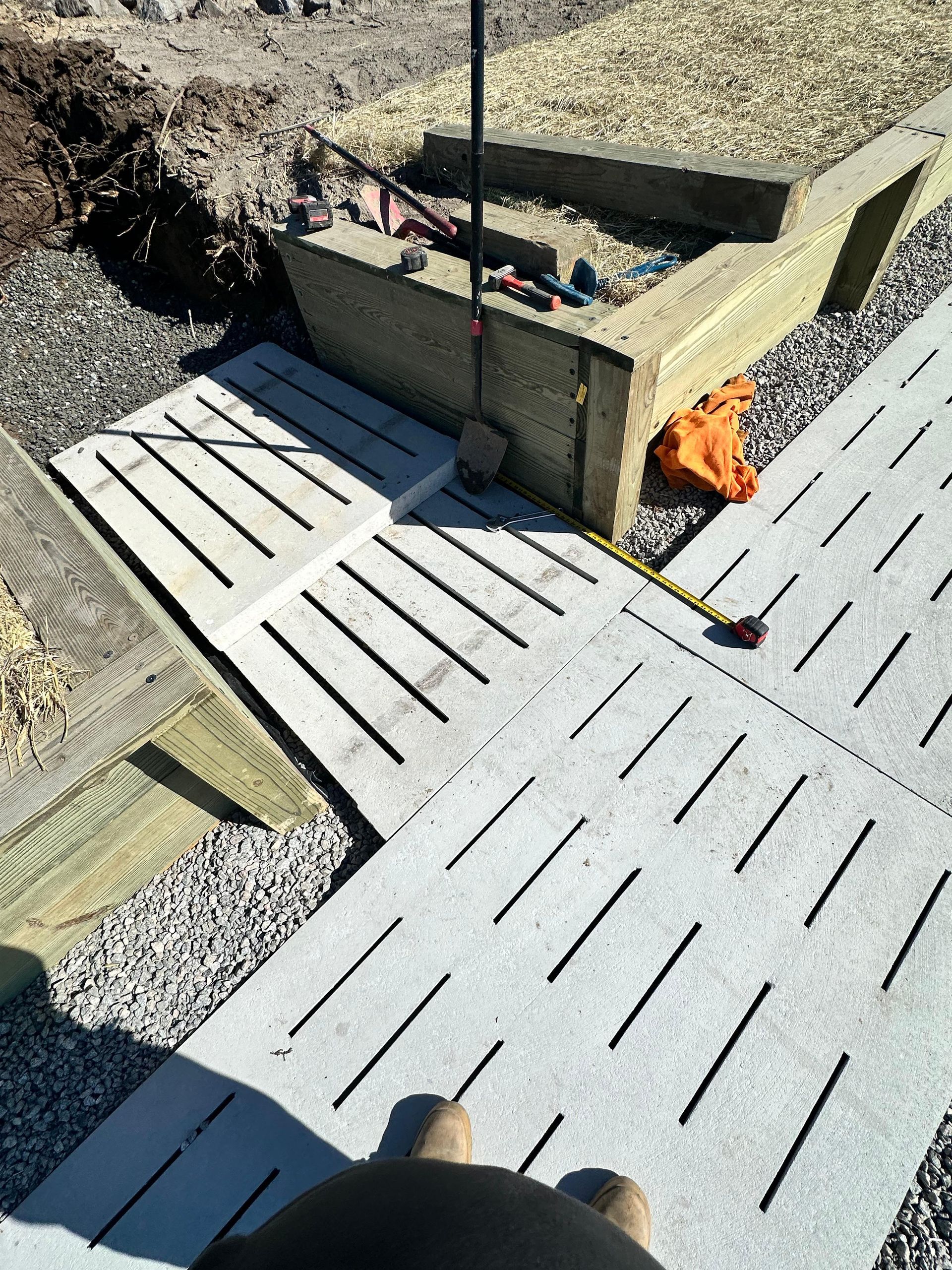 Concrete pavers with cut slots on a construction site; gravel, insulation, tools, and a person's feet.