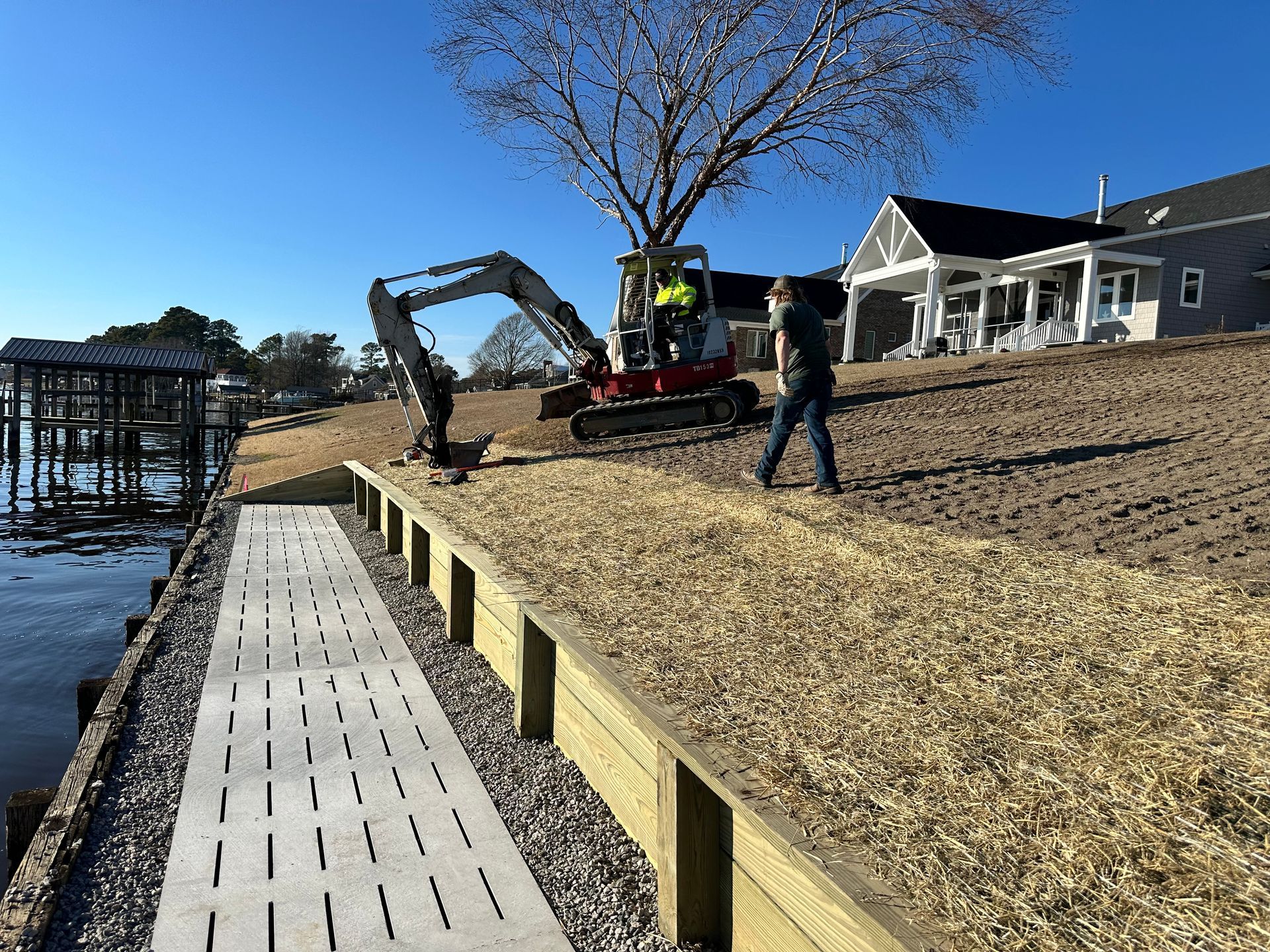 Construction on a waterfront with a person, excavator, wood retaining wall, and walkway.