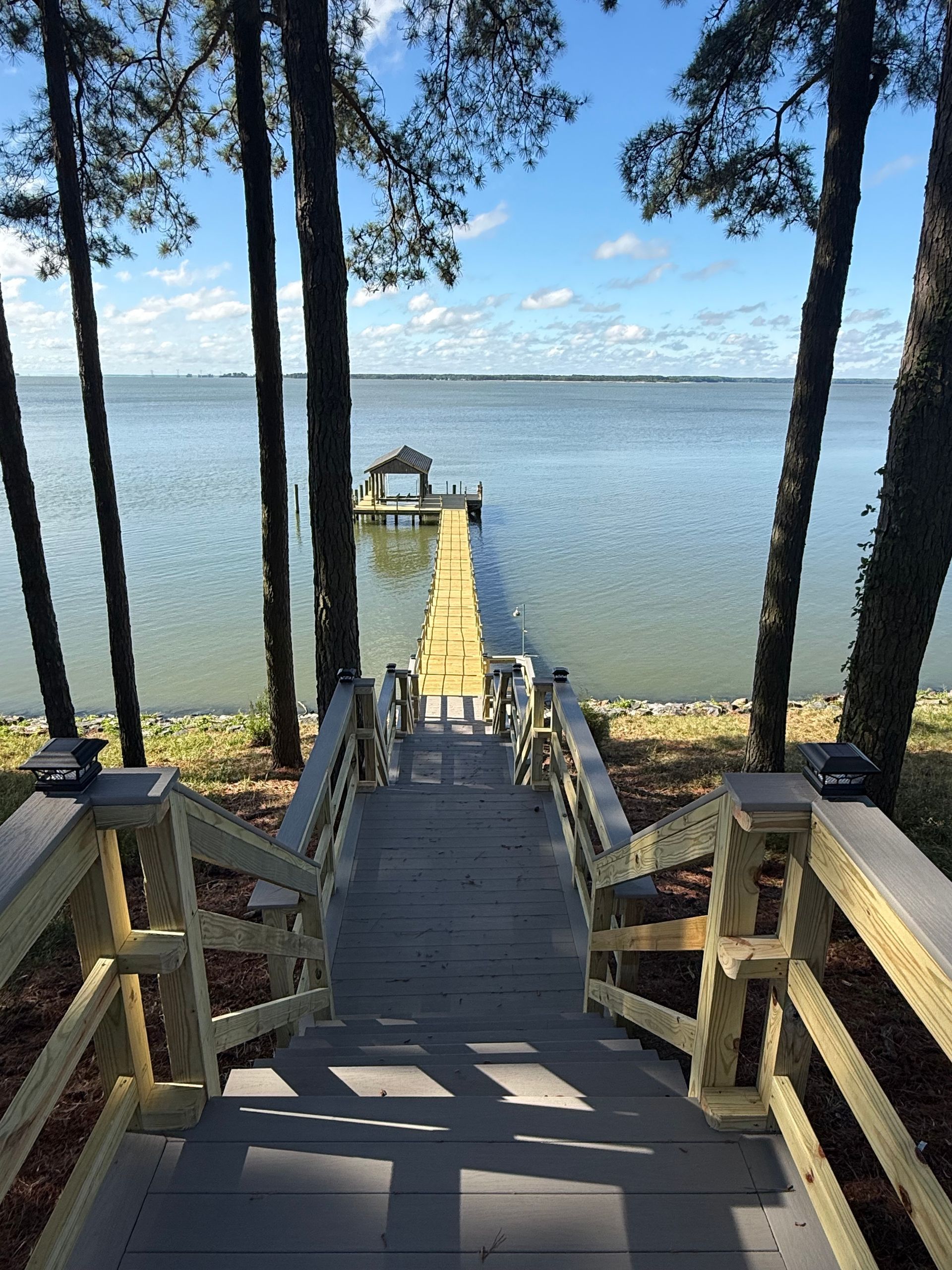Wooden staircase descending to a dock on a calm lake, framed by tall trees under a blue sky.