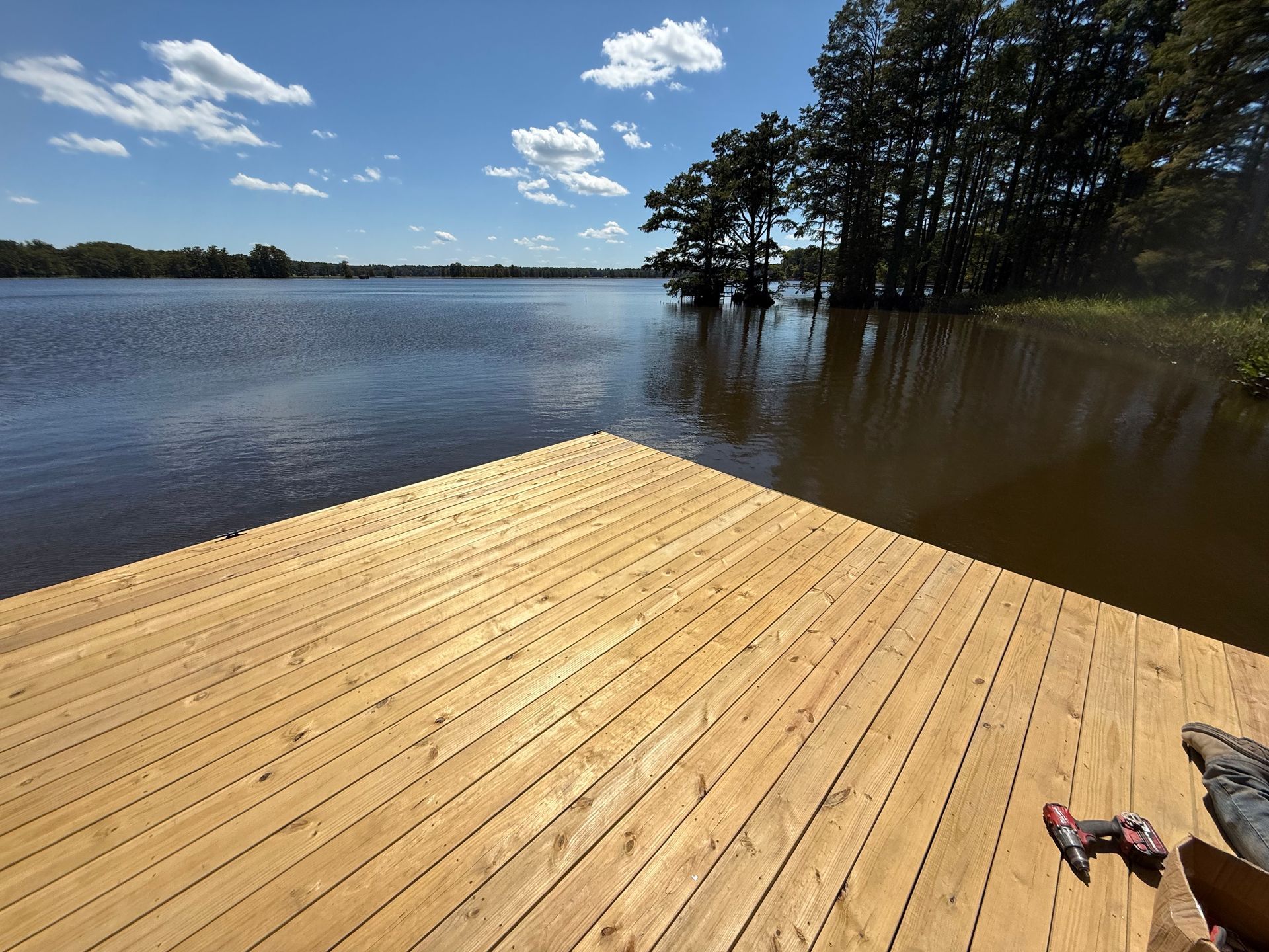 Wooden dock extending into a lake under a blue sky with clouds, trees in background.