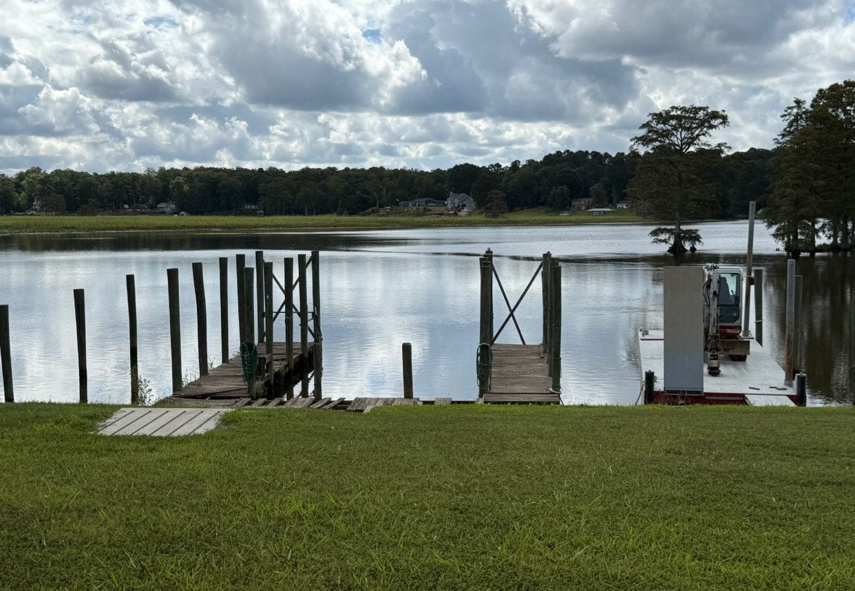 Wooden docks on a calm lake, lush green lawn in the foreground, cloudy sky above a distant tree line.