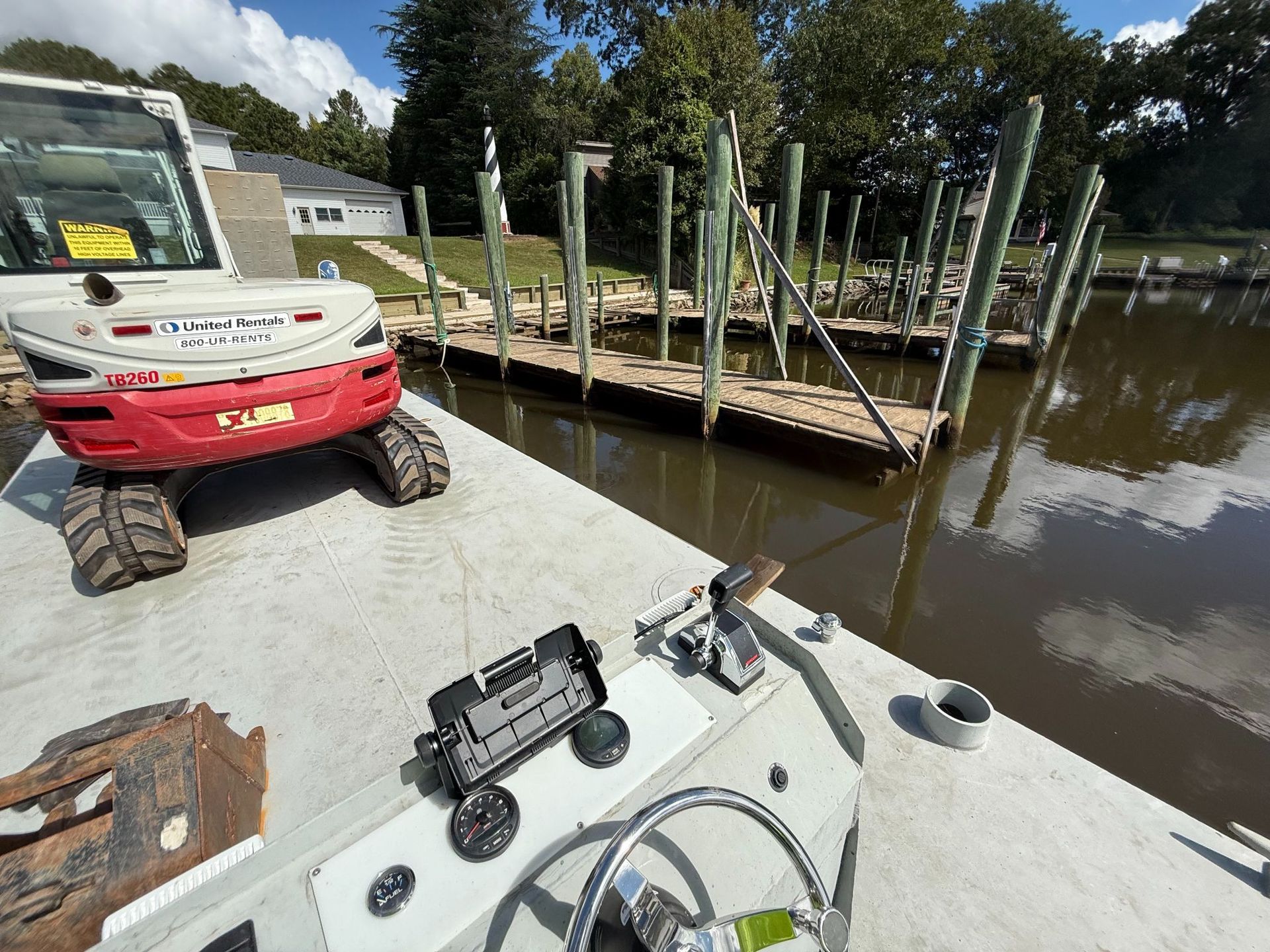 A construction scene on a dock with a small excavator, a barge, and water, under a blue sky.