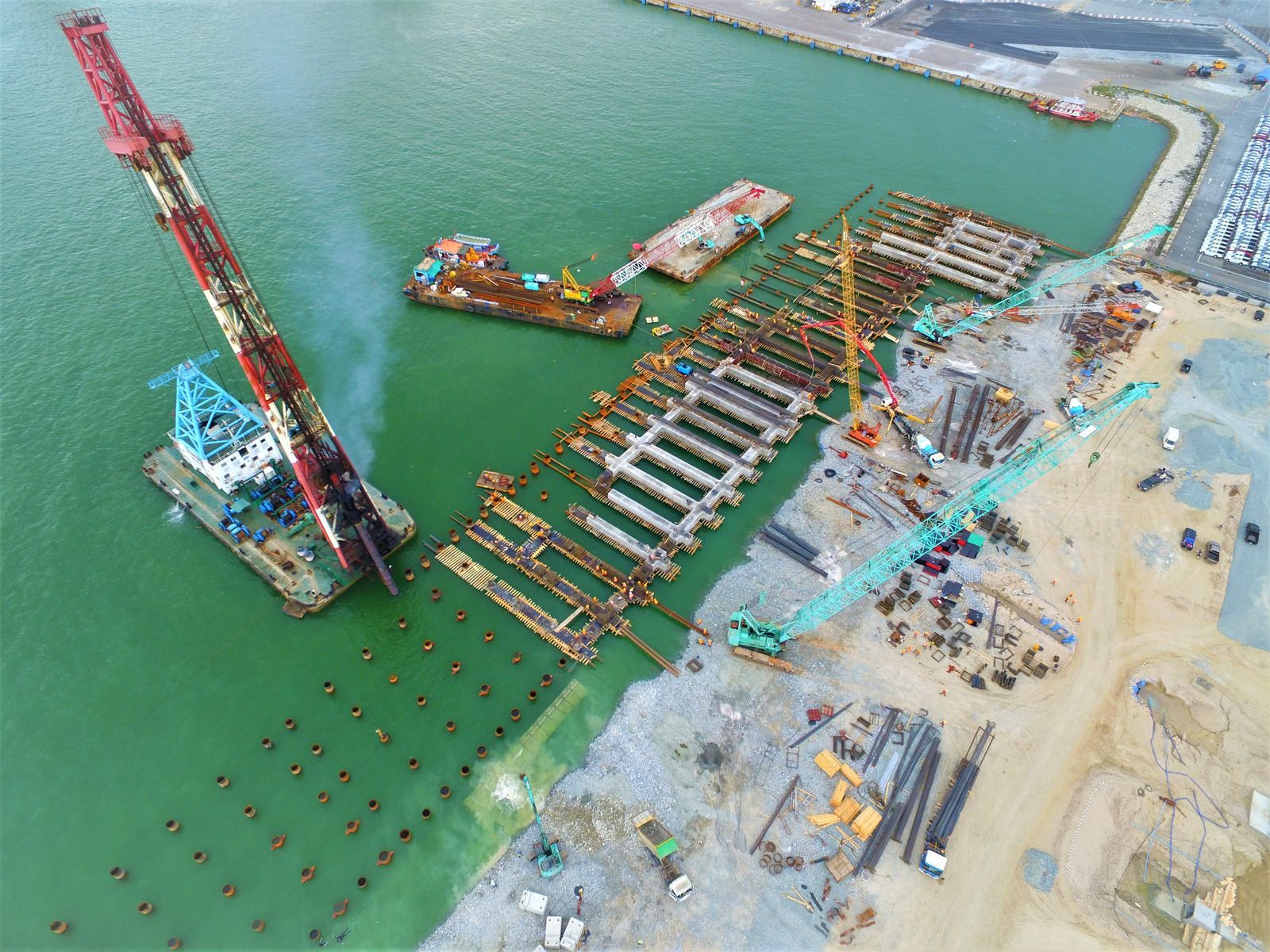 Aerial view of a construction site in water, with crane, docks, and machinery; green water and sandy shore.