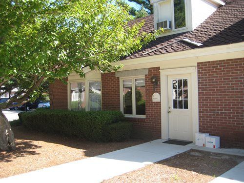 Brick building entrance with white door, windows, and trimmed hedges under a leafy tree.