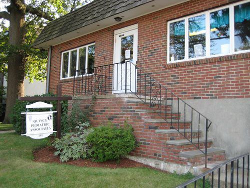 Brick house entrance with front steps, black railings, and a white sign by the lawn.