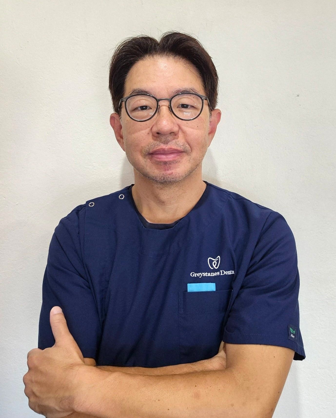 A man wearing glasses and a blue scrub top is standing in front of a white wall. A man wearing glasses and a blue scrub top is standing in front of a white wall.
