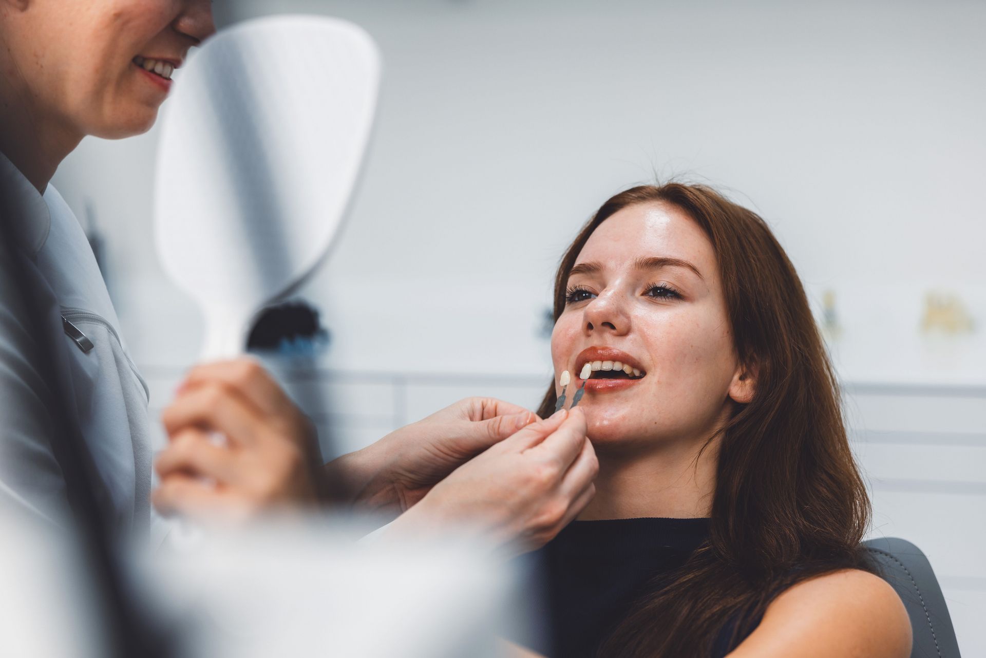 A dentist is fitting a dental veneer on a patient examining her smile in a handheld mirror.