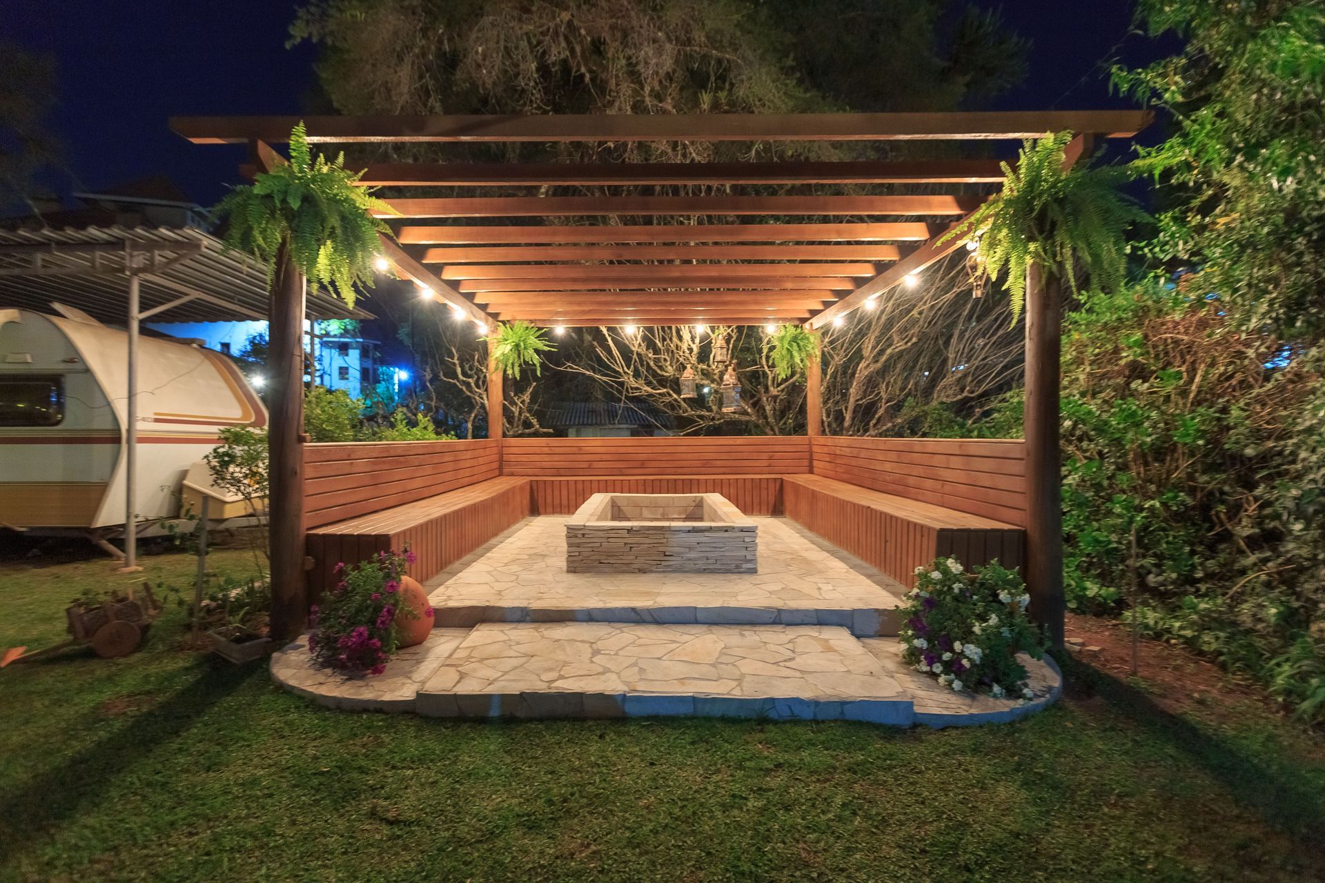 Covered backyard patio with a lit central fire pit, wooden pergola, and surrounding brick seating at night