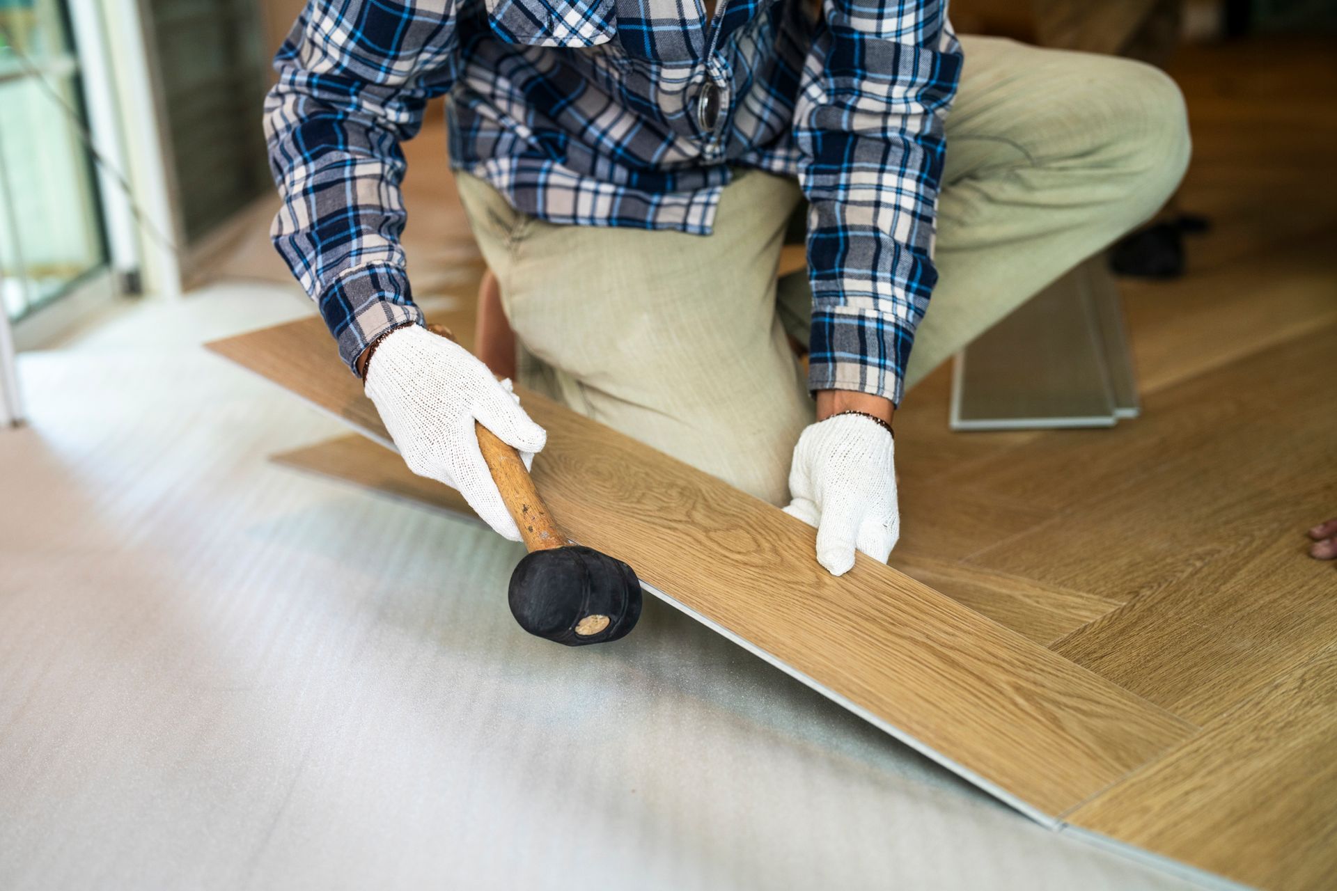 A man wearing gloves holds a hammer while working on hardwood flooring replacement in a home.