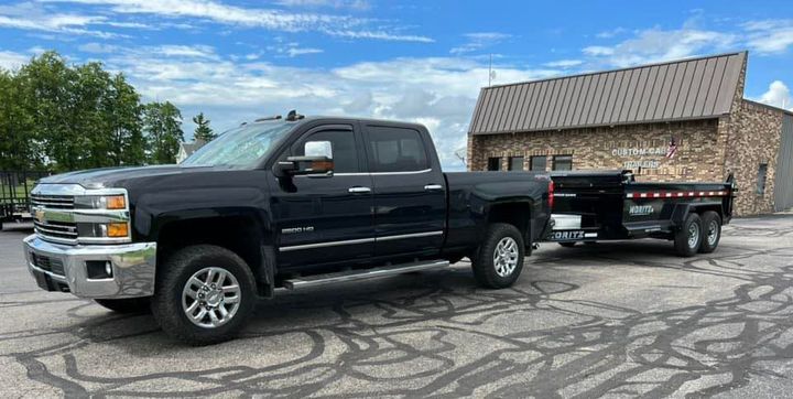 A black truck is towing a dump trailer in a parking lot.