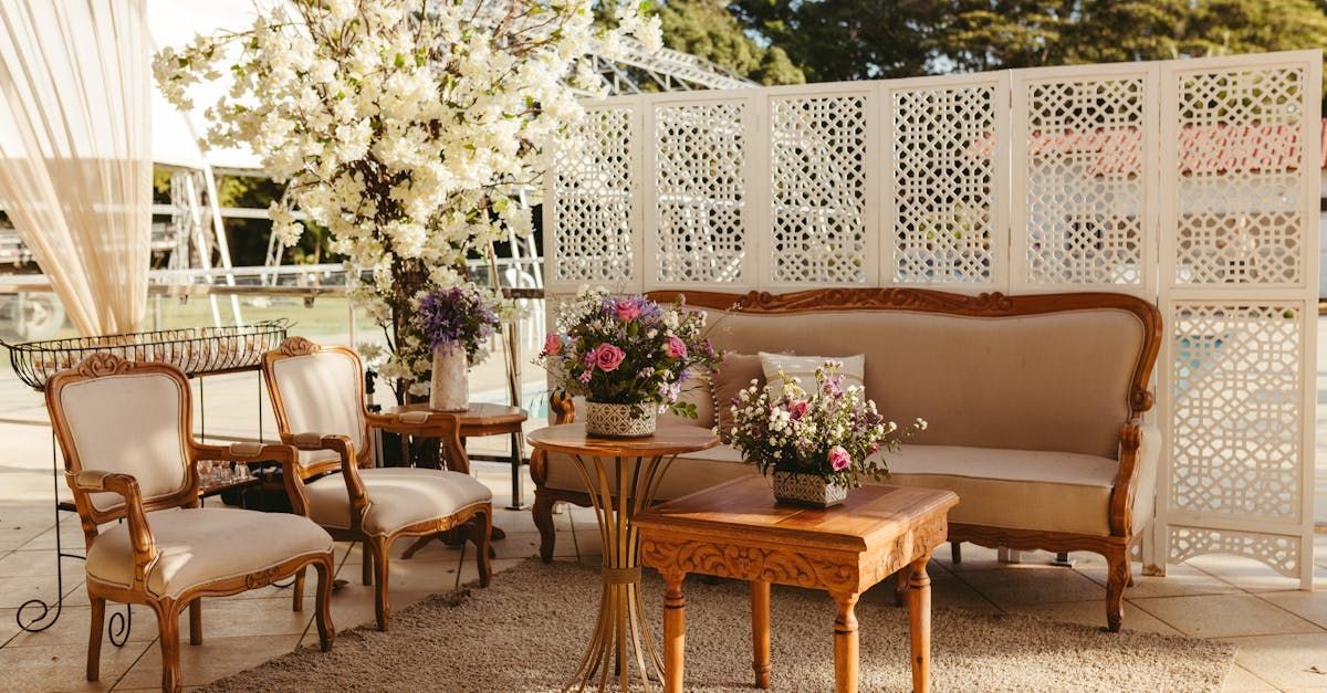 A couch , chairs , tables and vases of flowers are sitting in front of a white wall.