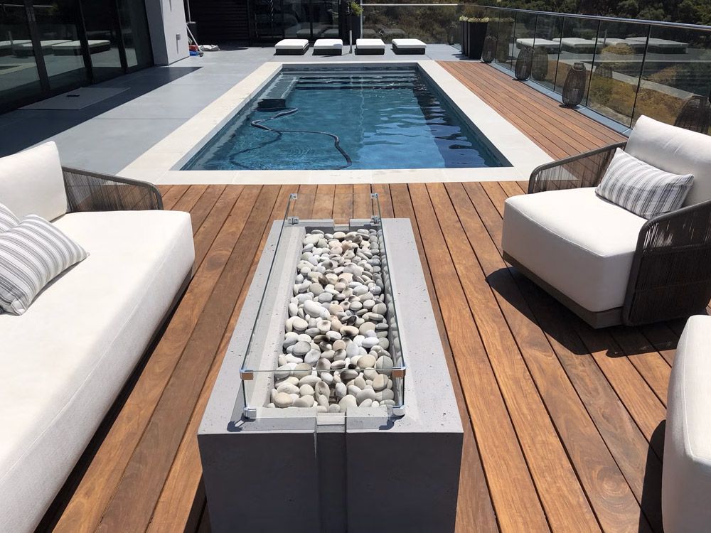Modern patio with a narrow pool, white sofas, and a central fire pit on a wooden deck.
