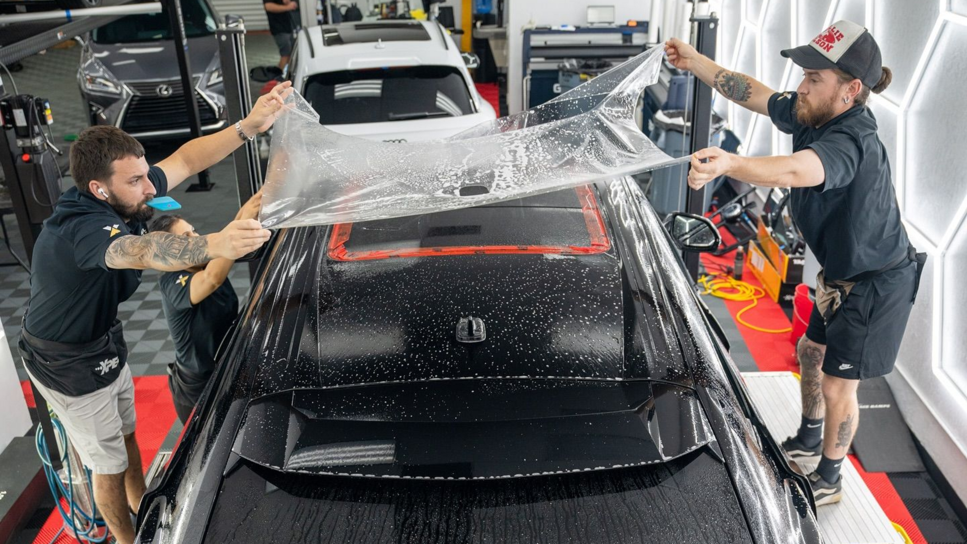 Two people wrapping clear film over a black car in a workshop