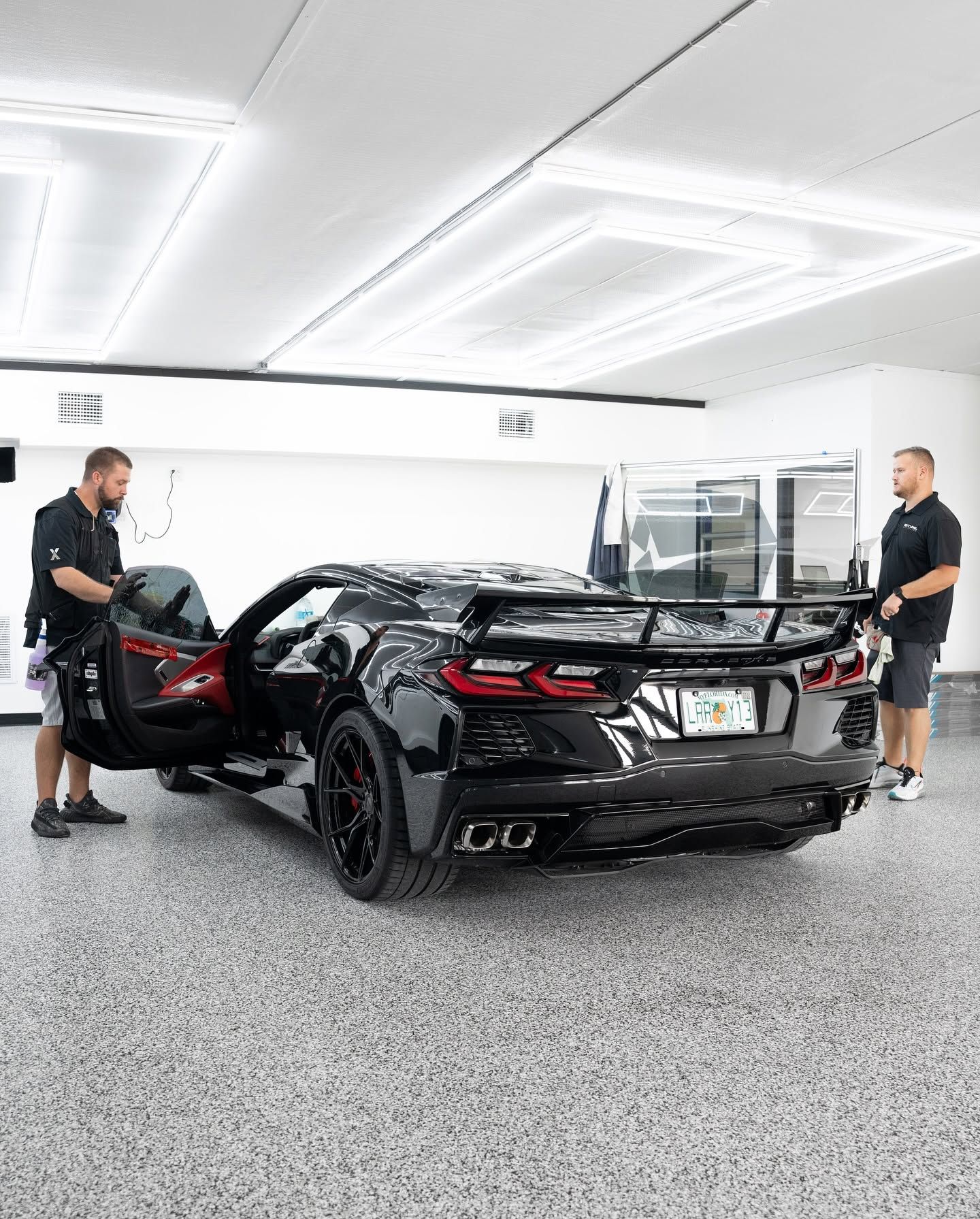 Two men stand by a black sports car in a bright showroom, one opening the driver’s door.