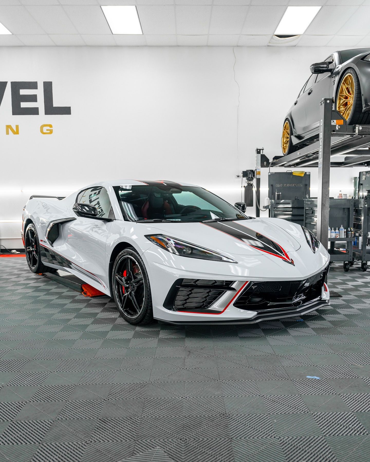 White Corvette with black racing stripes in a shop. Another car on a lift in the background.