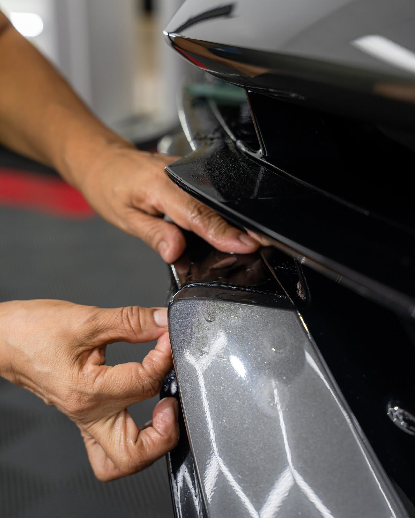 Hands adjusting a silver car bumper near a black front grille in a workshop