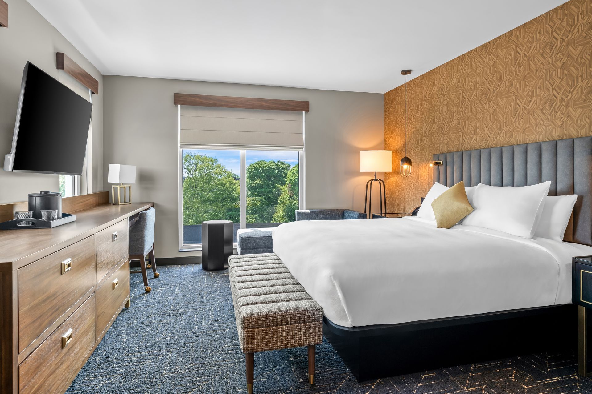 Modern hotel room with a king bed, bench, dresser, TV, lamp, and balcony view