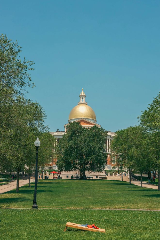 Massachusetts State House with golden dome, seen through trees and a grassy park under a blue sky.