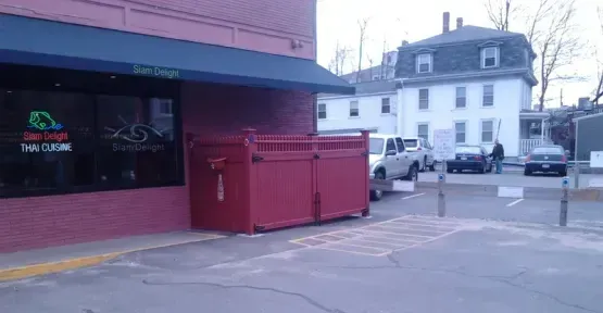 A red trash can is sitting in front of a building in a parking lot.