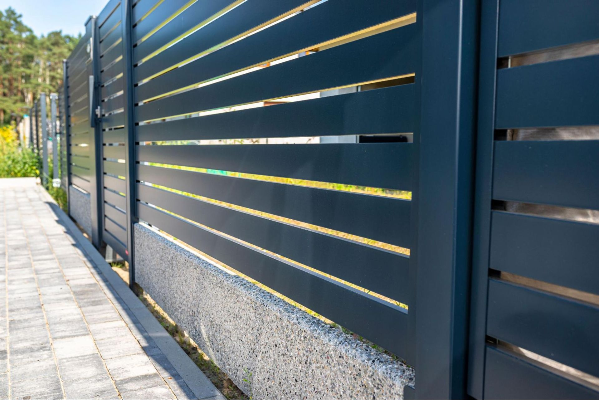 Dark gray horizontal slat fence with a stone base along a paved walkway. Trees are visible in the background.