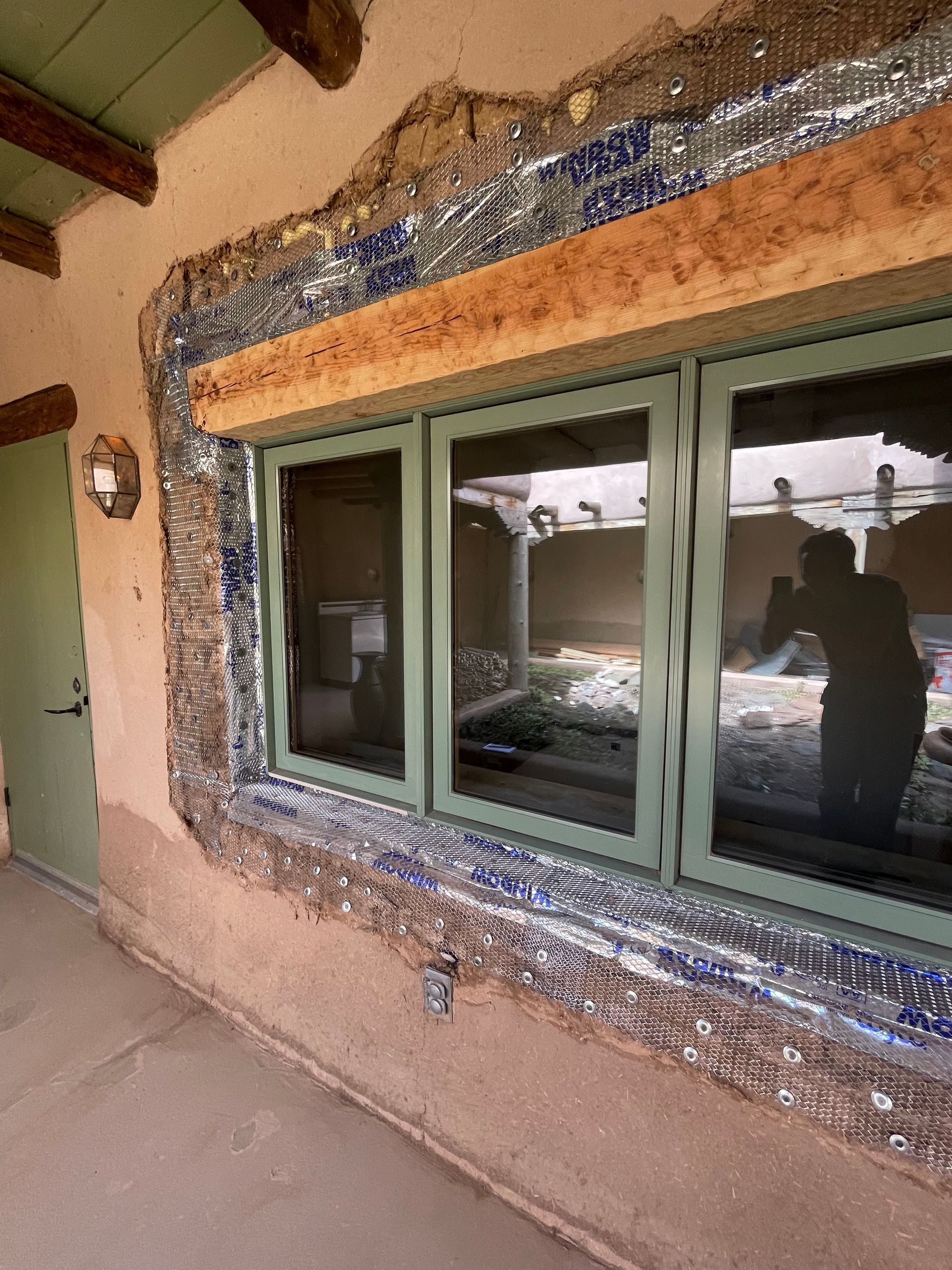 Exterior window with green trim, there is damaged adobe and exposed  metal lath  around the window