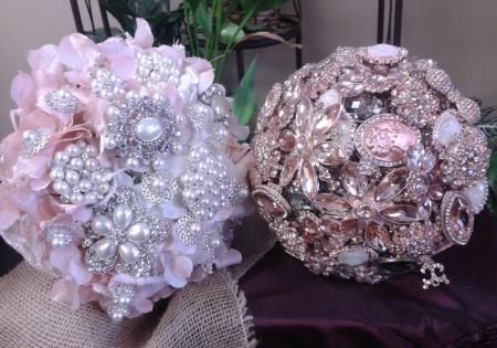 Two jeweled floral brooch bouquets, one pale pink and one rose gold, on a fabric surface