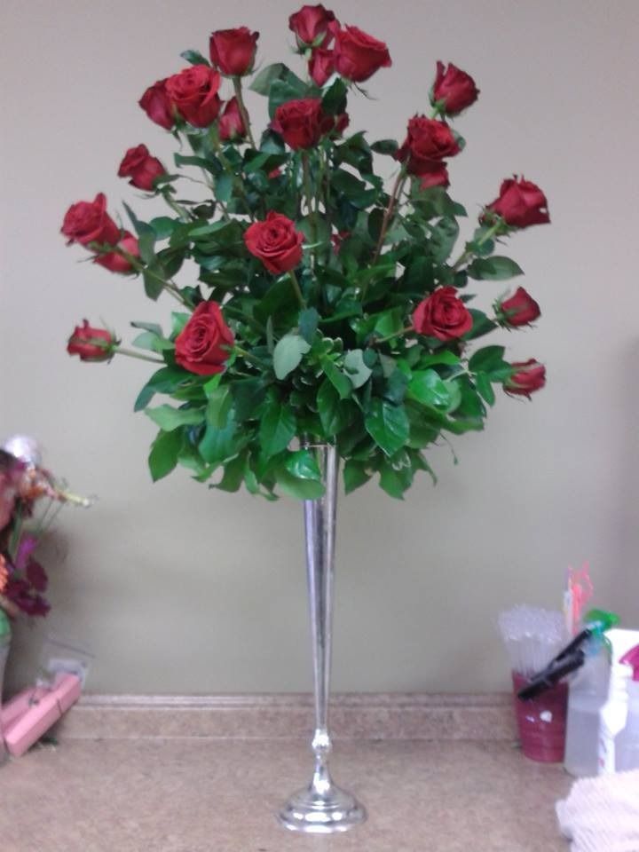 Red roses in a silver vase on a carpeted floor against a beige wall.