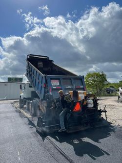 Dump truck on a gravel lot under a cloudy sky, with a front-mounted attachment and workers nearby.