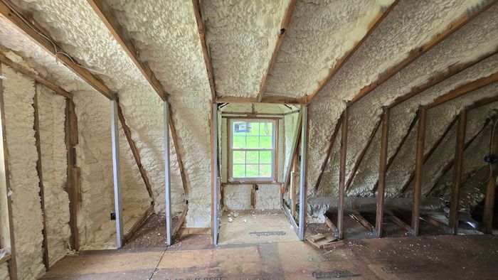 Attic with spray foam insulation, exposed framing, and a small window at the far end.